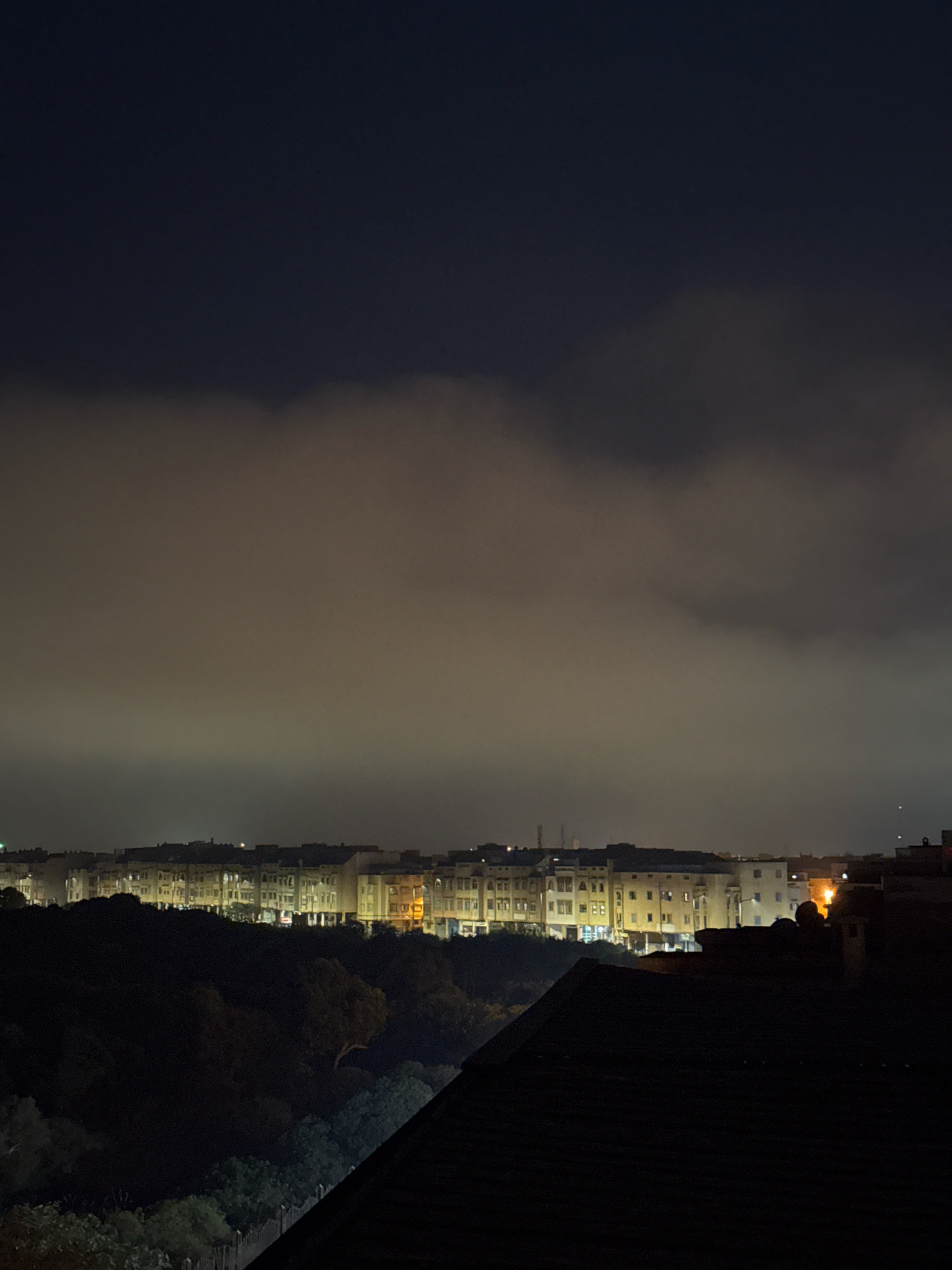 A nighttime aerial view captures a city skyline in Essaouira, Morocco. The sky is dark, with a band of illuminated clouds stretching across the middle ground. Below the clouds, a row of buildings is visible, their windows glowing with artificial light. In the foreground, the dark roof of a building dominates the lower portion of the image, with some trees visible on the left. The city appears densely populated, with numerous structures illuminated against the dark sky.