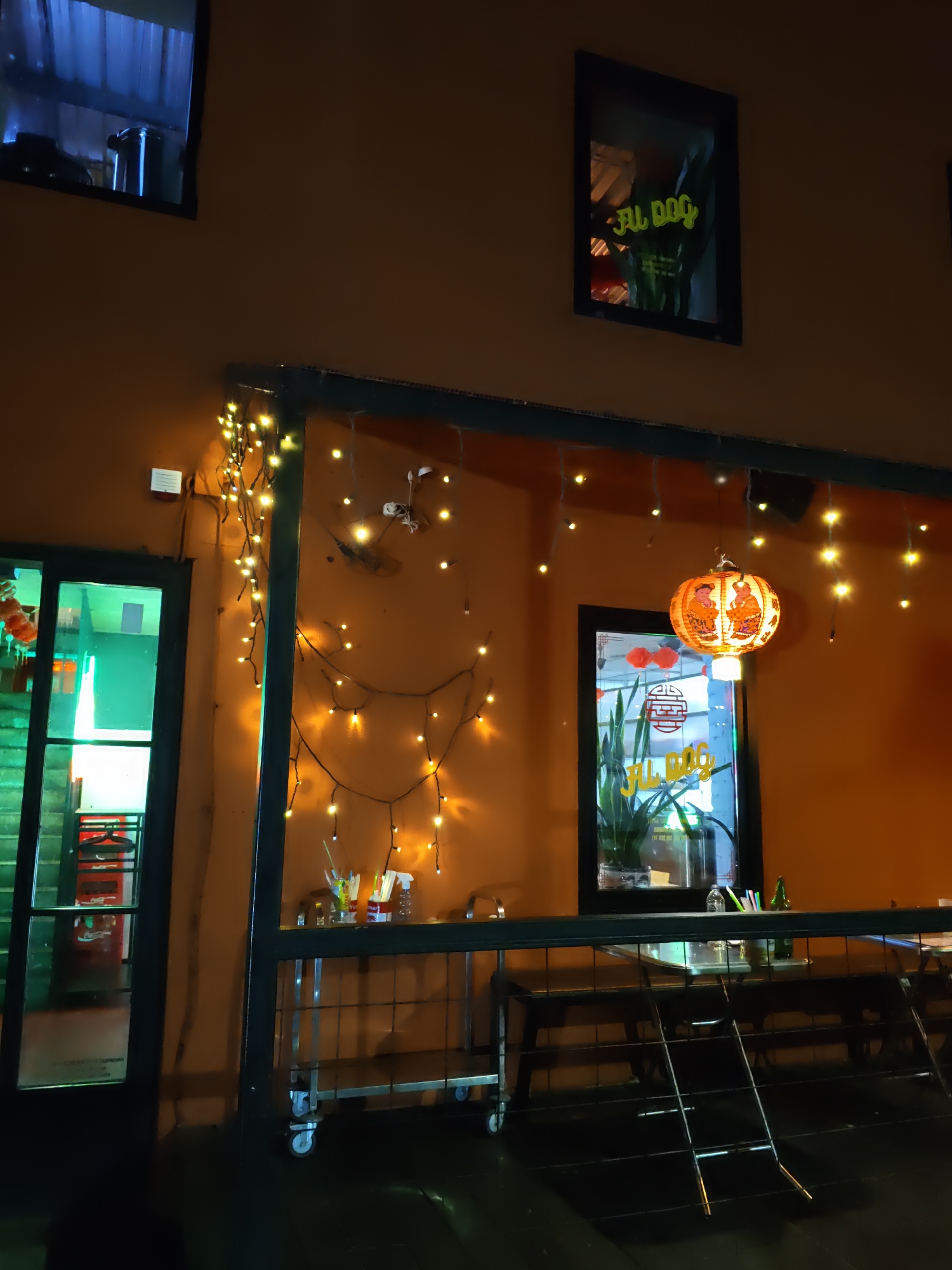 The image depicts the exterior of a restaurant at night. The building has an orange facade. There are two windows visible on the upper part of the facade; one on the left is dark with a blue interior light, and another on the right shows a neon sign reflected in it that reads "FU DOG". A string of yellow fairy lights is draped across the building's facade, creating a festive atmosphere. In the center of the image, a window is visible with a round, orange paper lantern hanging in front of it. The lantern is decorated with images of figures and Chinese characters. Inside this window, there are plants and a red circular emblem. To the left, there is a glass door with a green tint. Below the windows and the lantern, there is a railing and tables, suggesting an outdoor seating area. A metal cart with shelves is positioned near the entrance. A Coca-Cola cooler is visible through the glass door on the left. The lighting suggests it is nighttime in an urban environment. This scene is located in Rostov-on-Don, Russia.