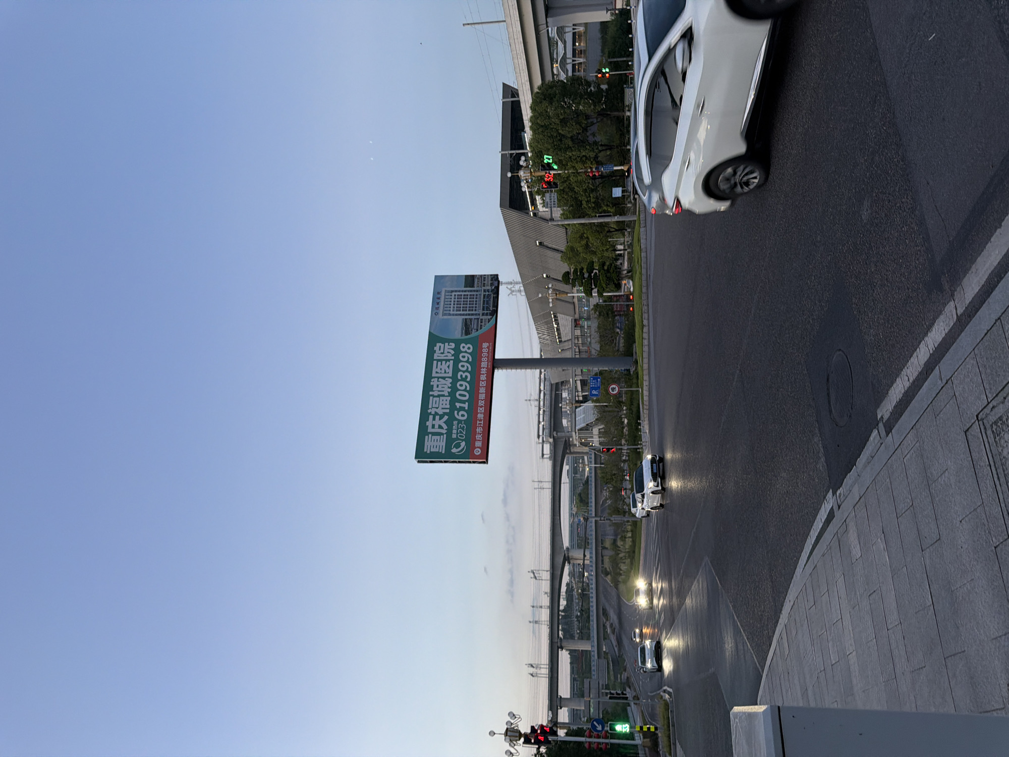 The image shows a city intersection in Jiangjin District, Chongqing, China, at dusk. A large billboard for Chongqing Fu Cheng Hospital is prominently displayed, featuring the phone number 023-61093998 and an image of a hospital building. Several cars are visible on the wet asphalt road, navigating through the intersection under the glow of streetlights and traffic signals. Overhead, a complex network of elevated roads and bridges, likely part of a highway or a high-speed rail line, dominates the background. The sky is a pale blue, transitioning into dusk, with a few wispy clouds. The overall scene captures a typical urban environment as evening sets in.