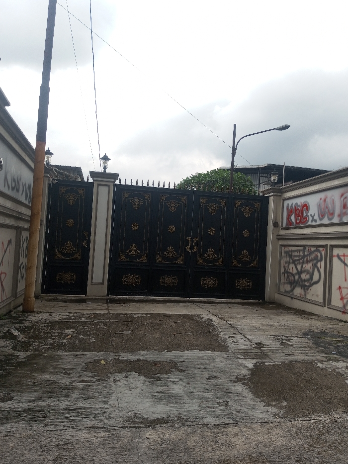 The image displays an exterior scene of a large property entrance in Bogor, Indonesia, under an overcast sky. A substantial, dark-colored metal gate, featuring ornate gold-colored scrollwork designs and pointed finials along its top, occupies the central view. The gate is supported by two light-colored pillars, each crowned with a decorative lantern-style light fixture. To the left, a light-colored perimeter wall shows black graffiti, including partially legible text "KISS XW" and "KB TO". To the right, a segmented perimeter wall is heavily marked with graffiti, notably "KBC XW" in red spray paint and multiple black scribbled tags; a single decorative light fixture is also mounted on its upper right section. A tall, dark utility pole stands vertically on the far left, partially wrapped in a lighter material near its base. Above the right side of the gate, a curved street lamp pole supports a horizontal luminaire. Thin utility lines are visible crossing the cloudy sky. The foreground comprises a grey concrete or asphalt driveway exhibiting extensive wear, stains, and patches of moisture. Green foliage from trees is visible behind the gate and walls. No people, vehicles, or specific actions are depicted.