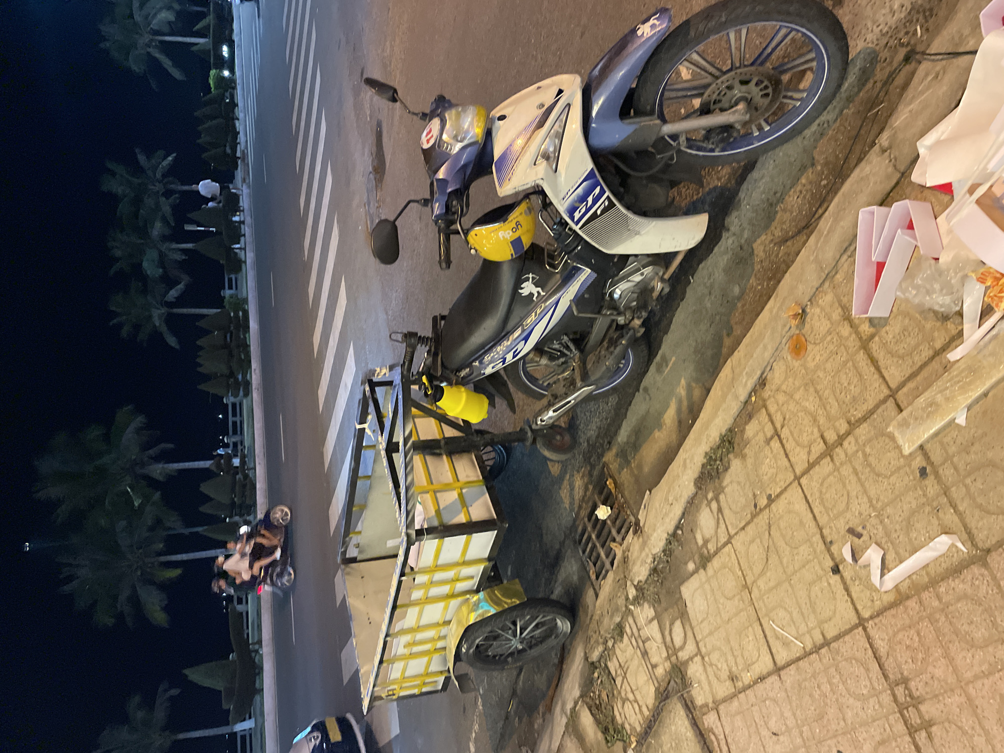 The image shows a motorized tricycle parked on a paved sidewalk next to a street. The tricycle is hauling a large, yellow cargo box. The scene is set at night, with streetlights and the dark sky suggesting artificial lighting. Palm trees line the street in the background, indicating a tropical or subtropical location. A few people are visible on the street, either walking or riding other motorcycles. There are also some white and red packaging materials scattered on the sidewalk near the tricycle. The overall impression is of a street scene in an urban or semi-urban area in Vietnam at night.
There is some text visible on the motorcycle, but it is not legible in the image. The text on the yellow helmet reads "R p oy". The motorcycle itself has a blue and white color scheme with some graphic designs. The tricycle appears to be a common mode of transport for goods in the region. The lighting conditions suggest it is after sunset. The presence of palm trees and the style of the motorcycle are consistent with a location in Southeast Asia, specifically Nha Trang, Vietnam.