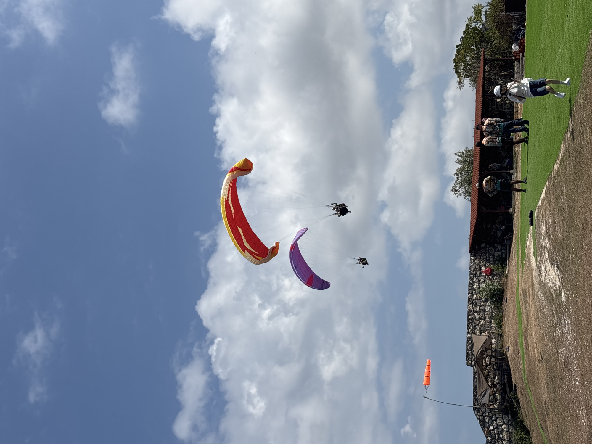 This image shows a paragliding launch site in Kuta Selatan, Indonesia. The sky is blue with scattered white clouds. Three paragliders are in the air. The largest paraglider, in the upper right, is red and yellow. Below and to the left of it, a purple paraglider is visible. In the distance, a smaller paraglider is also in the air. On the ground, a group of people are gathered near a building with a red roof. A windsock is visible on a pole to the left of the building. The ground is covered in dry grass with patches of green.