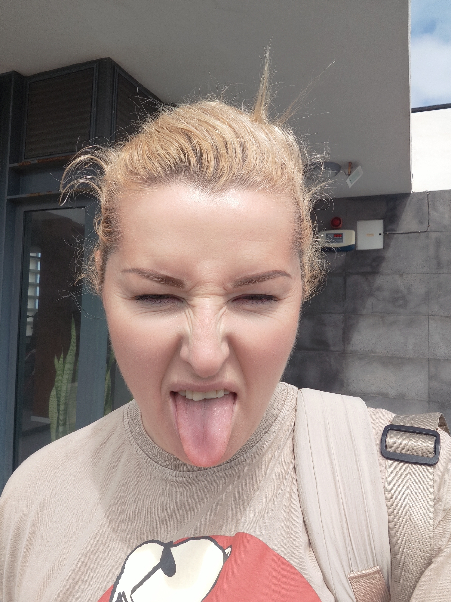 The image depicts a fair-skinned woman with blonde hair pulled up in a messy bun. She is sticking her tongue out and squinting her eyes, suggesting a playful or silly expression. She is wearing a beige t-shirt with a graphic design on the front, and a backpack strap is visible over her shoulder. The background shows an urban environment with a building exterior, including a window, a concrete wall, and what appears to be an alarm system panel. Based on the description of the location, the image was taken in Ribeira Grande, Portugal.