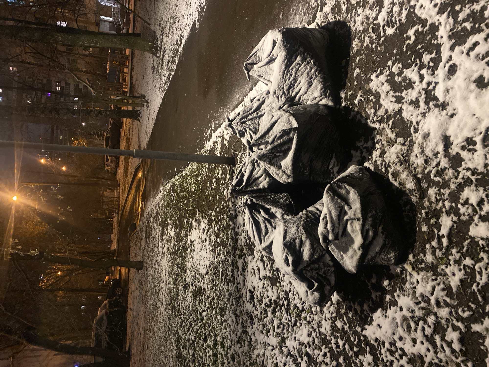 The image depicts a nighttime scene in what appears to be a park or residential area, with snow covering the ground and various objects. Four large black trash bags covered in snow are piled near a lamppost. To the left, bare trees and a building with lit windows are visible, suggesting a city environment. The lighting is artificial, with the lamppost and other distant lights illuminating the scene. Based on the presence of snow and architecture, the location is identified as Moscow, Russia.