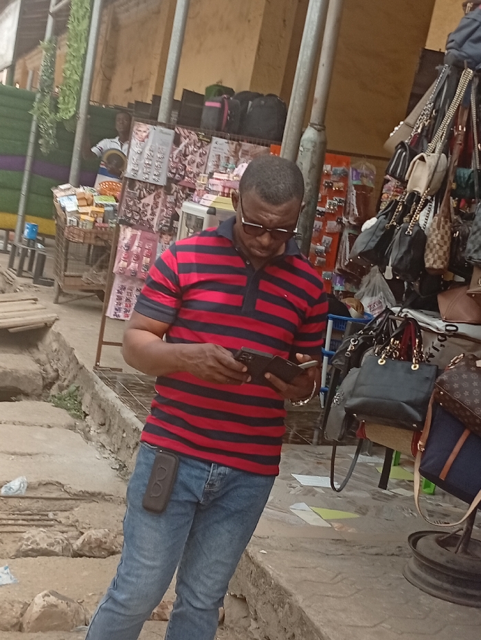 The image captures an outdoor market scene, likely during the daytime, with a focus on a man in the foreground. The overall atmosphere suggests a bustling commercial area, possibly a street market or a series of informal stalls.
In the center of the frame, a man with a dark complexion and short, closely cropped dark hair is prominent. He is wearing dark-framed sunglasses, a short-sleeved polo shirt with horizontal red and black stripes, and light blue denim jeans. His head is tilted downwards, and he is intensely focused on a dark-colored smartphone or similar device held open in both hands at waist height. A black, rectangular phone holster or case is clipped to the right side of his jeans. He also appears to be wearing a bracelet on his left wrist.
Behind the man, the background reveals multiple market stalls. To the left, a display rack holds numerous pairs of sunglasses, earrings, and other small accessories, suggesting a fashion accessory stall. Another individual, lighter-skinned and wearing a white shirt, can be partially seen peering from behind this stall in the upper left background. Further left, there's an area with green and purple colored panels or walls, adding a splash of color to the otherwise earthen tones.
To the right of the central figure, another stall is visible, heavily adorned with various handbags and purses of different sizes, colors, and styles, hanging from racks. One notable handbag features a distinctive monogram pattern, possibly indicating a designer brand replica. The ground in the foreground is uneven, appearing to be a mixture of dirt and rough concrete or paving stones, with some white plastic litter scattered about. Tall, unpainted metal poles support the overhead structures of the market stalls. The lighting is diffused, suggesting it's not direct midday sun, possibly late morning or early afternoon, with some areas appearing to be in partial shade. No clear, readable text or signage is visible in the image.