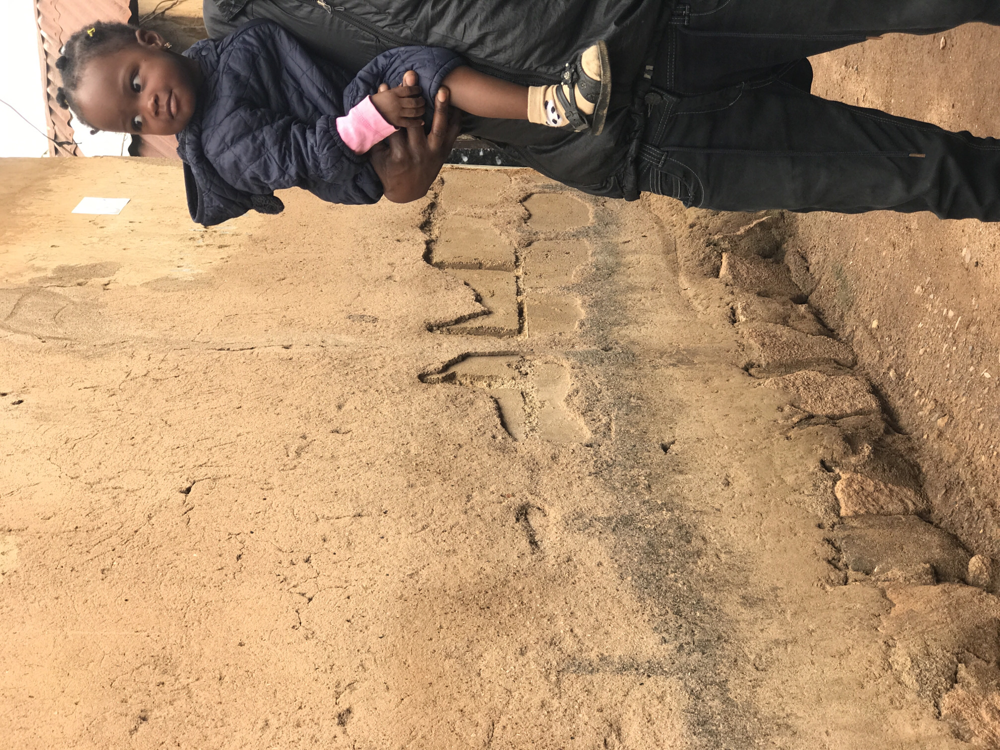 The image depicts a close-up view of a person carrying a young child in Kabong, Nigeria. The background is dominated by a textured, light brown wall, possibly made of mud or stucco, with some darker, weathered marks and visible structural imperfections. The ground in the foreground is sandy and reddish-brown, with small pebbles.
The child, who appears to be a toddler, is facing to the right and looking away from the camera. The child is wearing a dark blue, quilted hooded jacket with pink sleeves peeking out. The child's feet are clad in tan socks with a panda design and black sandals. The child is being held by a person whose body is partially visible on the right side of the frame. This person is wearing a black puffer vest over a dark shirt and dark blue jeans. Their hands are holding the child. The person's skin tone appears to be dark. The lighting suggests an overcast day.