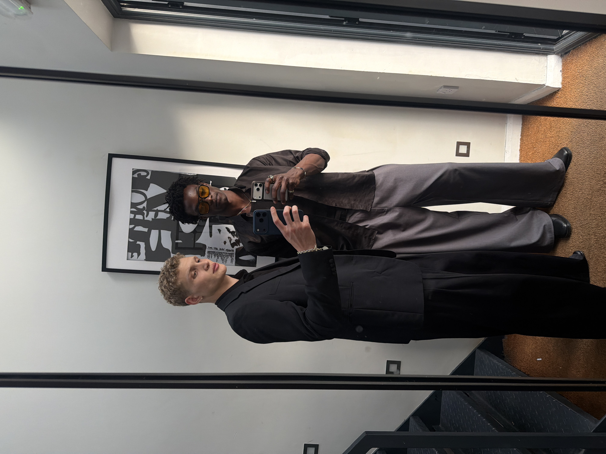 Two young adult males are visible in a full-length mirror selfie, positioned in a modern indoor setting identified as being in the city of London, United Kingdom, evidenced by the presence of a British Standard BS 1363 electrical outlet on the right wall.
The subject on the left, with light skin and short, curly blonde hair, wears a black suit jacket over a black collared shirt, paired with black wide-leg trousers and black shoes. A silver stud earring is visible on his left ear, and a silver chain bracelet adorns his left wrist. He holds a dark blue smartphone in his right hand, angled towards the mirror.
The subject on the right, with dark skin and dark, curly hair, wears yellow-tinted aviator-style sunglasses. He is dressed in an unbuttoned brown long-sleeved collared shirt over a dark undershirt, grey wide-leg trousers, and black shoes. Multiple silver necklaces are visible around his neck, and silver rings adorn his fingers. He holds a light-colored smartphone in his left hand, also angled towards the mirror.
The background consists of a plain white wall. To the left of the subjects, a black metal industrial-style staircase with diamond plate treads descends. Above and behind the subjects, a black-framed artwork with black and white abstract or text-based design is mounted on the wall. The floor is covered with a textured, medium brown carpet. The mirror itself is framed in black. To the far right, a vertical black frame suggests a window or glass door.