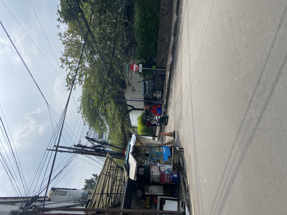 The scene is a street corner in Bandung, Indonesia, located at -6.923 latitude and 107.61 longitude. The street is paved and appears to be relatively empty, with a single motorcycle parked at the corner. There are a few people walking in the distance and a small shop on the corner. The street is lined with trees, some of which have branches reaching over the street. There are many power lines overhead. The sky is partly cloudy.