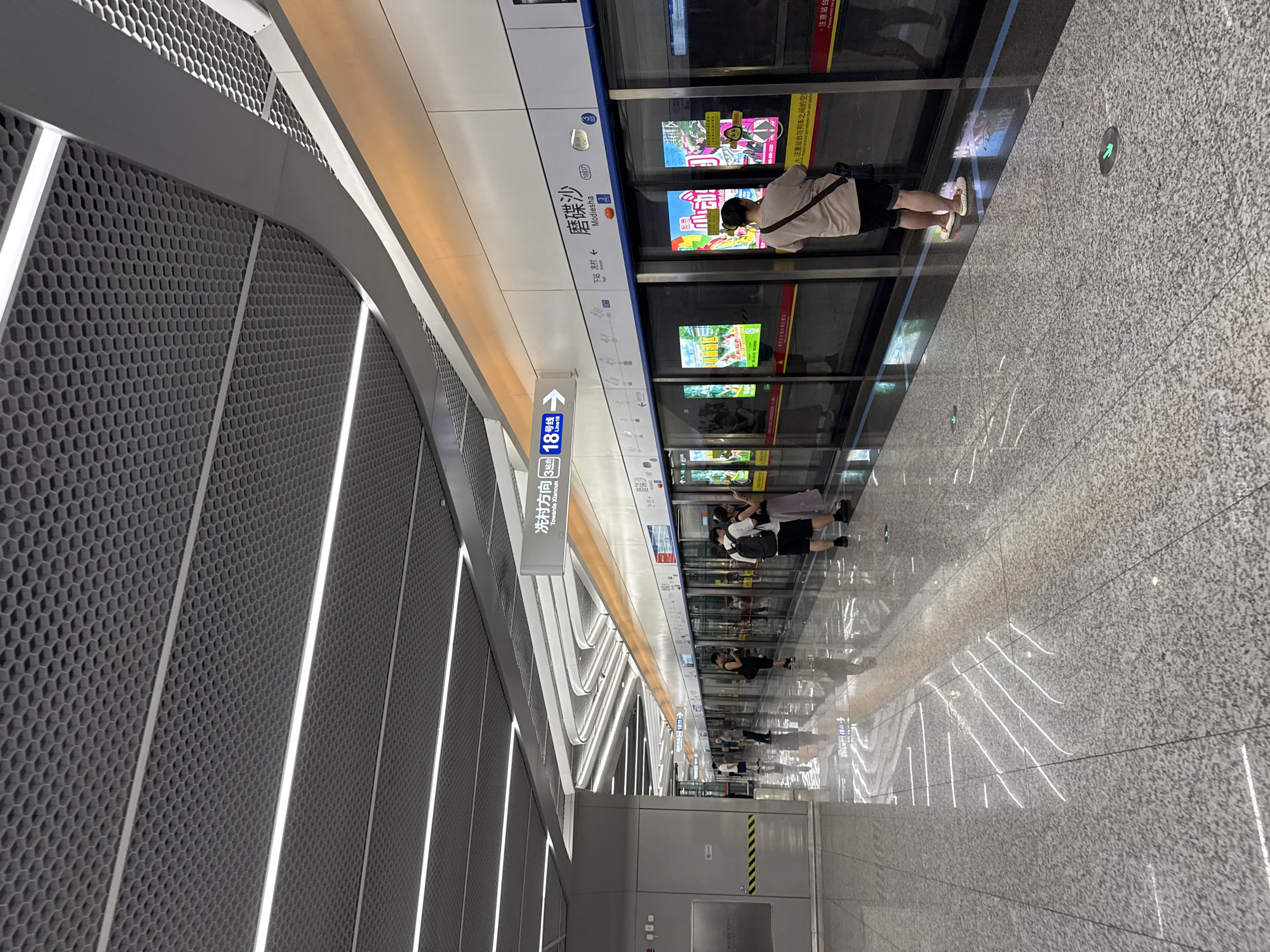 The image depicts a modern subway station platform in Haizhu District, China. The platform is clean and well-lit, with a polished granite floor that reflects the overhead lighting. The ceiling features a honeycomb-patterned acoustic paneling with integrated LED strip lighting that creates a futuristic aesthetic.
A subway train is stopped at the platform, with its doors open. Several passengers are visible: some are waiting to board, while others are already on the platform. The passengers are dressed casually, and some are carrying backpacks.
Signage in both Chinese and English indicates the direction of travel ("Towards Xiancun") and the line number ("Line 18"). The station name appears to be "Modiesha" (磨碟沙). Advertisements are displayed on the train doors. The overall atmosphere is one of efficiency and modern urban infrastructure.