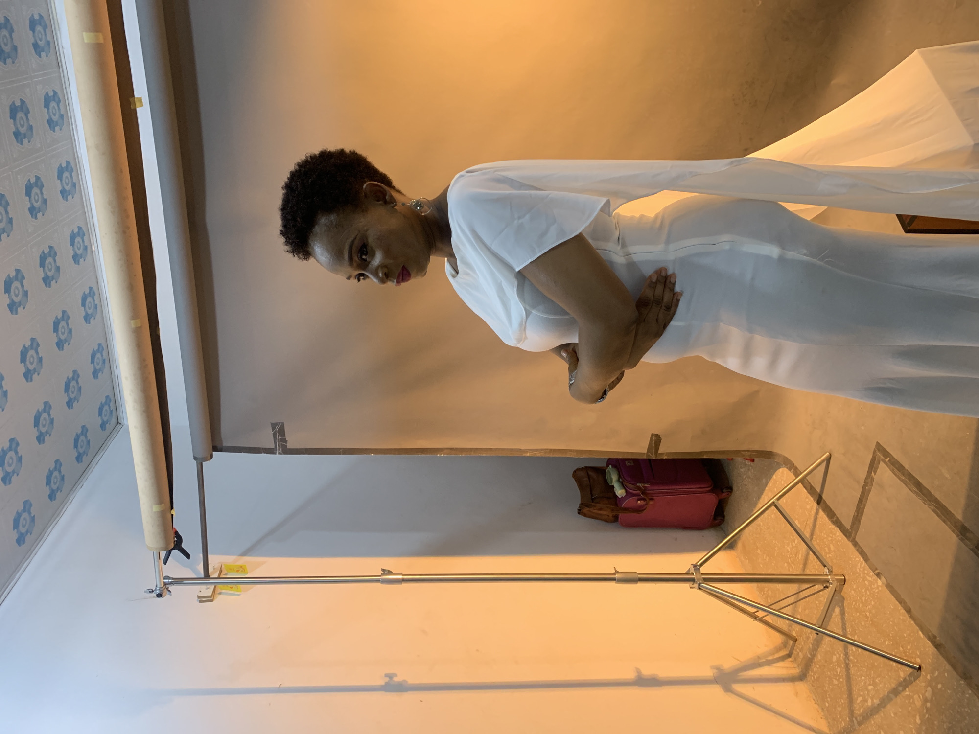 A woman with short, dark curly hair is posing indoors in front of a photography backdrop. She wears a white dress with draped sleeves and has her arms crossed. The backdrop is a neutral beige color, and there is a lighting setup visible in the image. The environment includes photographic equipment, such as a stand, and what appears to be a suitcase, possibly belonging to a photographer or other related personnel. There is no discernible information that would allow the specification of whether the location is in Gura Top, Nigeria.
