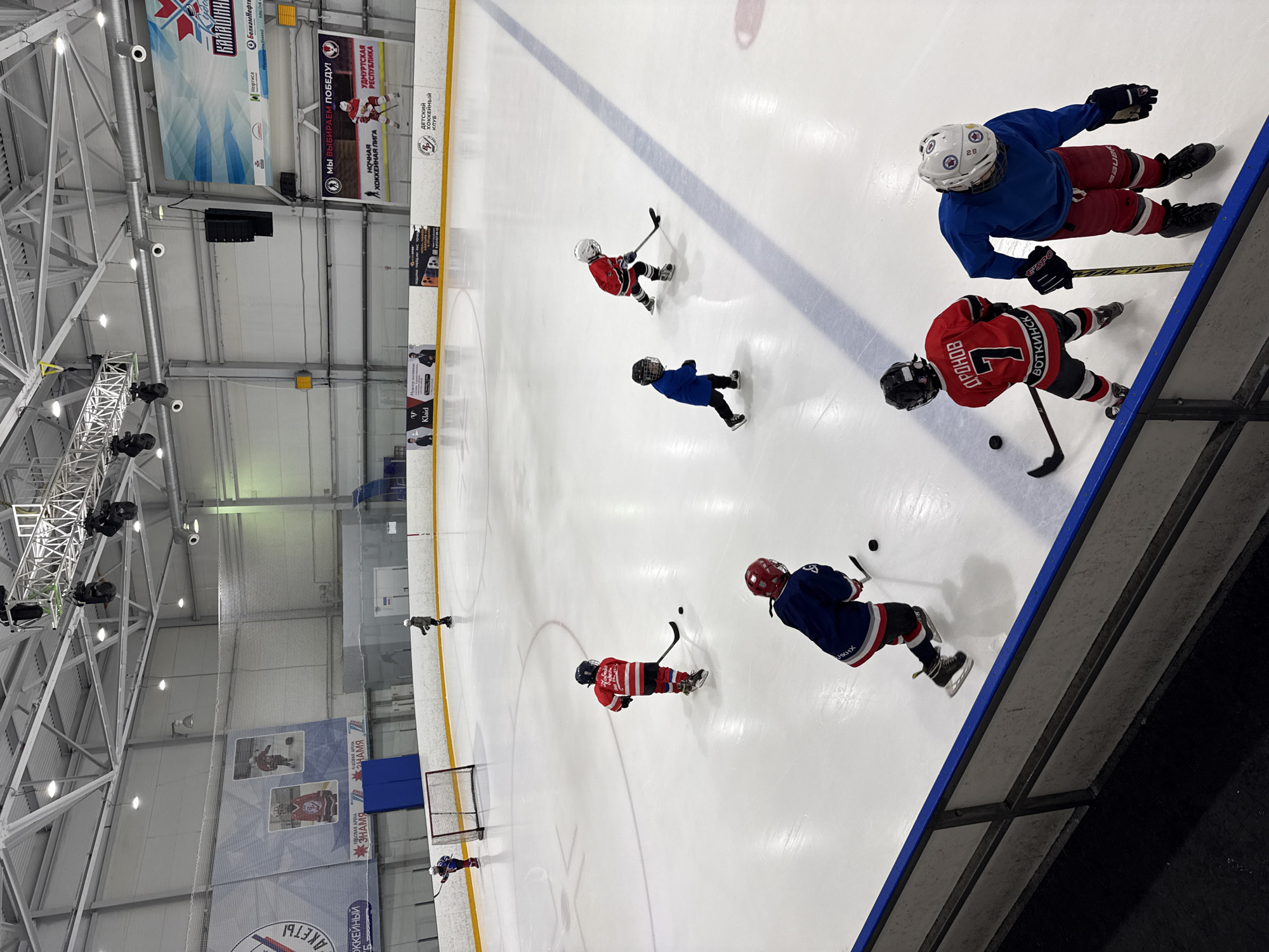 An indoor ice hockey rink in Votkinsk, Russia, is shown from a high-angle perspective. Six young hockey players in full gear are on the ice. Four players are actively engaged in skating or practicing, while two are further away, possibly waiting their turn. The rink has standard markings and a goal net is visible in the background. Large banners with text in Russian are displayed along the arena walls. The ceiling features exposed metal structures and lighting fixtures. The overall scene depicts a youth hockey training session.