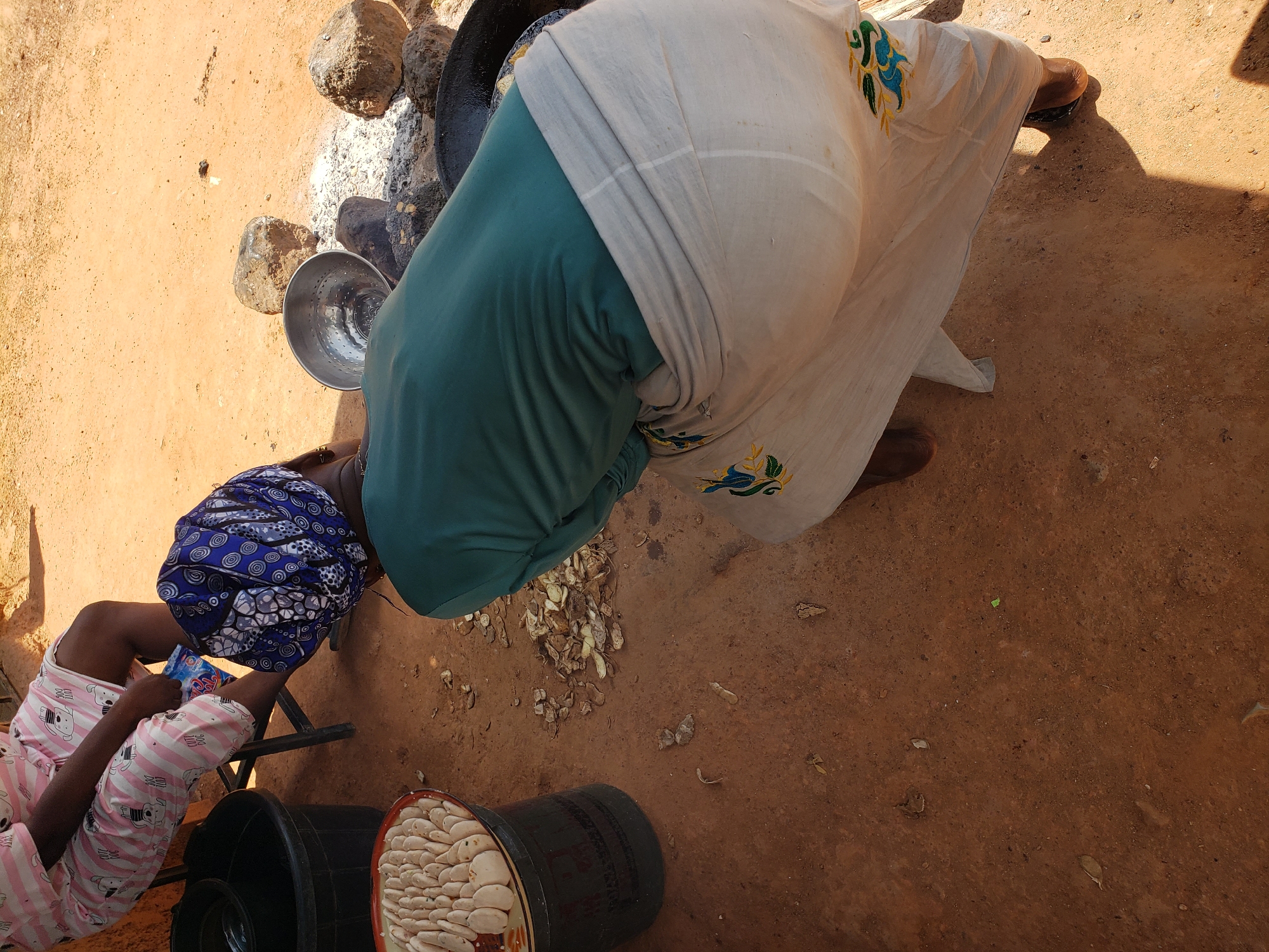 In Narayi, Nigeria, an outdoor scene unfolds on a dusty, reddish-brown earth ground under bright sunlight. The image captures two individuals engaged in an activity, possibly food preparation. In the foreground, a person wearing a teal long-sleeved shirt and a light-colored, flowing skirt embroidered with blue and green patterns is bent over a black cooking pot, partially obscured from view. The skirt is wrapped around their waist and falls to their feet, which are shod in dark sandals. Beside them, a large metal bowl with a perforated interior sits near rocks and ashes, suggesting an open fire setup.
To the left, a second individual is seated on a black folding chair, wearing a pink and white striped outfit adorned with dog graphics. Their legs are crossed, and they are holding a blue packet. In front of this person, a black plastic basin is visible, and to its left, a round, shallow metal tray is filled with numerous small, pale, oval-shaped objects, possibly food items such as dough balls or dumplings, arranged in a neat pattern. Scattered on the ground are dried leaves and small pieces of debris. The background shows more of the dusty ground and a hint of an orange-colored wall.