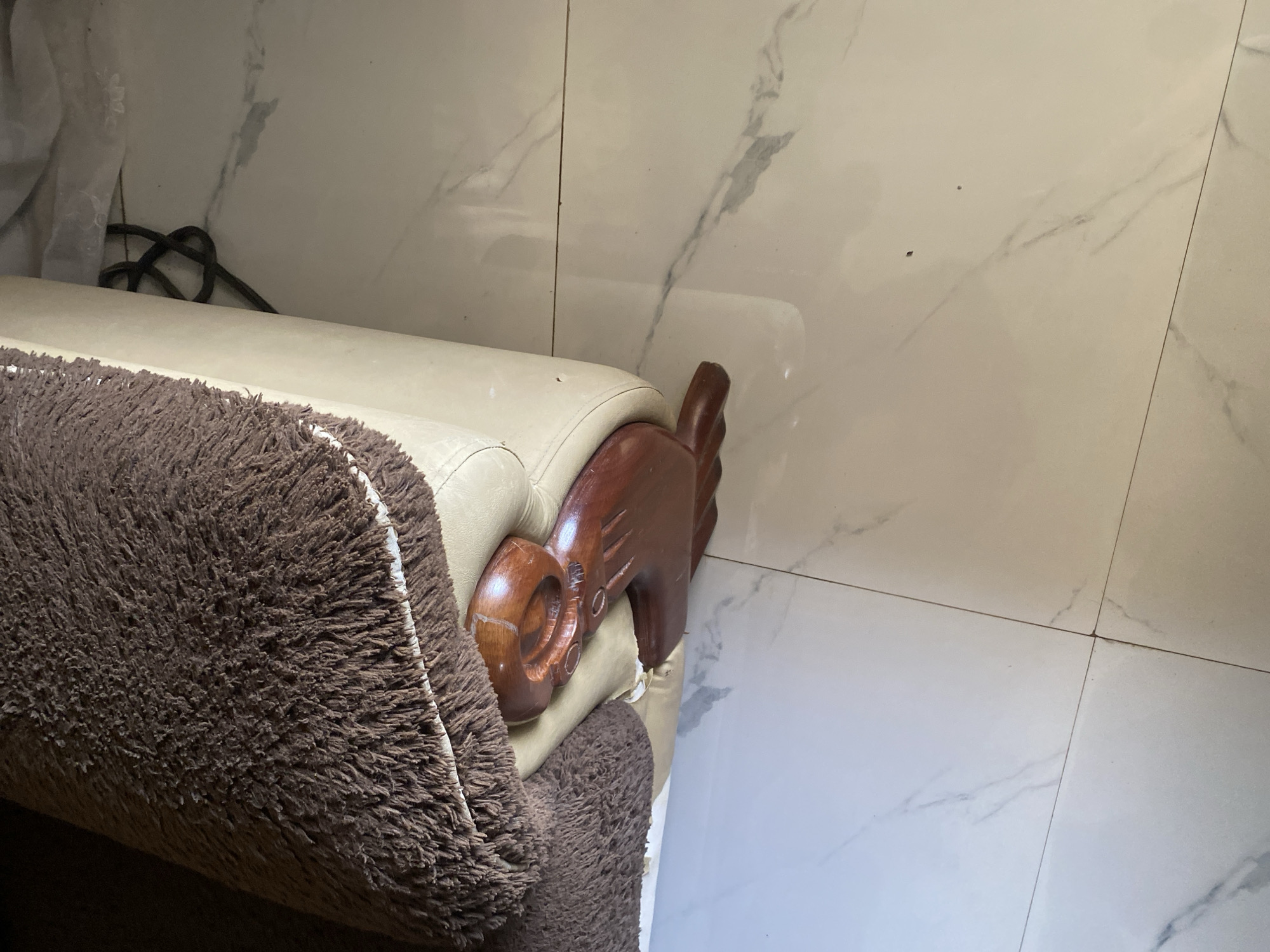 A brown fluffy blanket is draped over the side of a cream-colored sofa. The sofa has a carved wooden leg that is elaborately shaped. The floor is tiled with a white marble-like pattern. The scene is indoors and appears to be a living area. The location is Jauro Yinu, Nigeria.