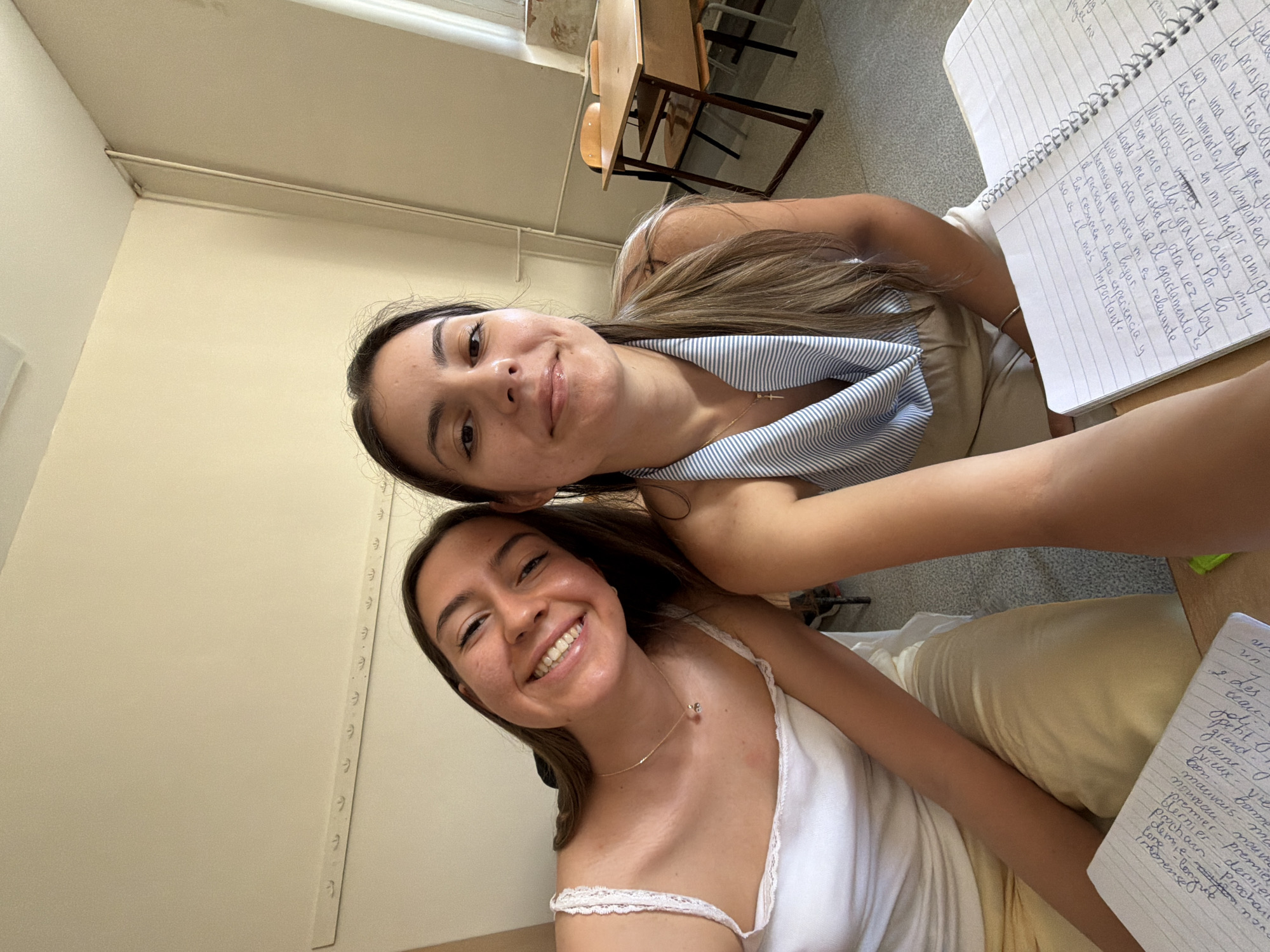 The image shows two young women taking a selfie in what appears to be a classroom or study area. The woman on the left is wearing a white tank top with lace trim and yellow pants. She has a necklace with a small pendant. The woman on the right is wearing a striped top and beige pants. Both women are smiling at the camera. A notebook with handwritten text is visible in the foreground, along with parts of desks and chairs in the background, suggesting an educational setting. The overall lighting indicates it is daytime. The wall color is a light beige. There is a metal bar with hooks along the top of the wall. The presence of desks and chairs, along with the overall ambiance, points to an indoor educational environment.
