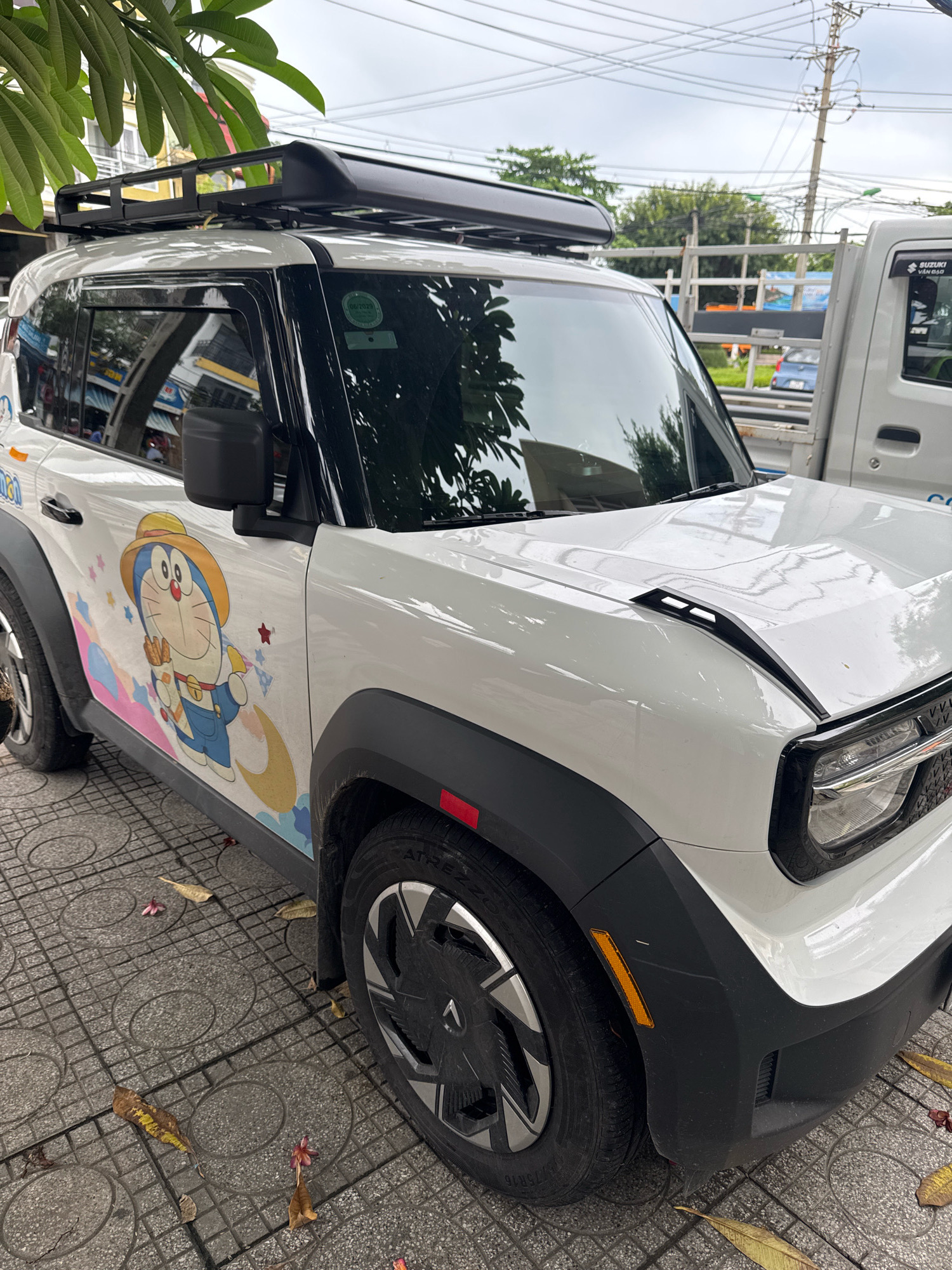 A white SUV with a cartoon character decal on its side is parked on a tiled sidewalk next to a street. The SUV has a black roof rack and black fender flares. Fallen leaves are scattered on the sidewalk. In the background, a white truck and buildings can be seen. The scene appears to be in Nha Trang, Vietnam, as indicated by the presence of a white truck with Vietnamese lettering on its side door which reads "SUZUKI VĂN ĐẢO".