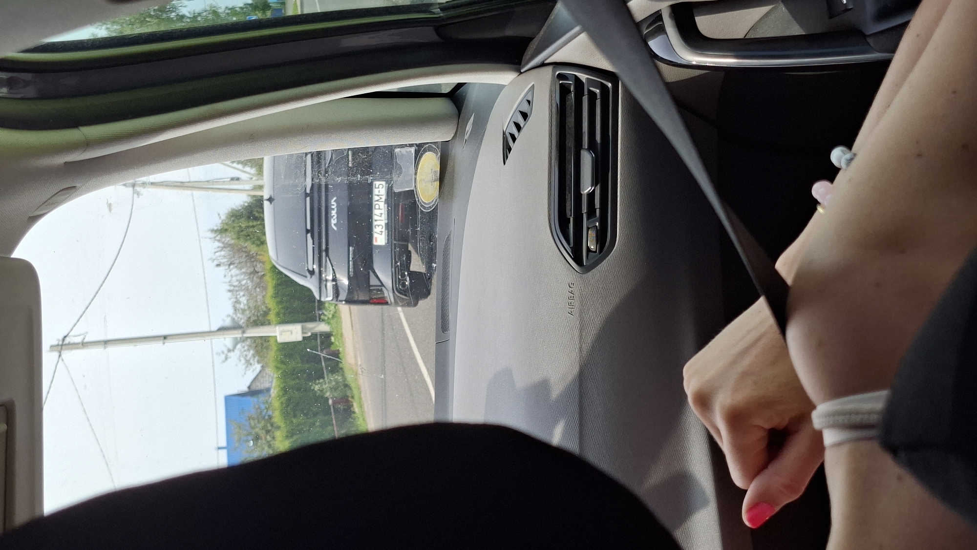 The image captures a view from inside a car, looking forward towards the road. In the foreground, a woman's hands are visible, one with bright pink nail polish resting on a seatbelt. The dashboard of the car is also visible, with an "AIRBAG" label and an air vent. Further ahead, a black car is driving on a paved road. The license plate of the car reads "4314PM-5". The background shows a rural landscape with trees, a green hedge, and a building with a blue roof. Utility poles and wires are present along the roadside. The sky is overcast. Based on the license plate, the location is likely Myadzyel, Belarus.