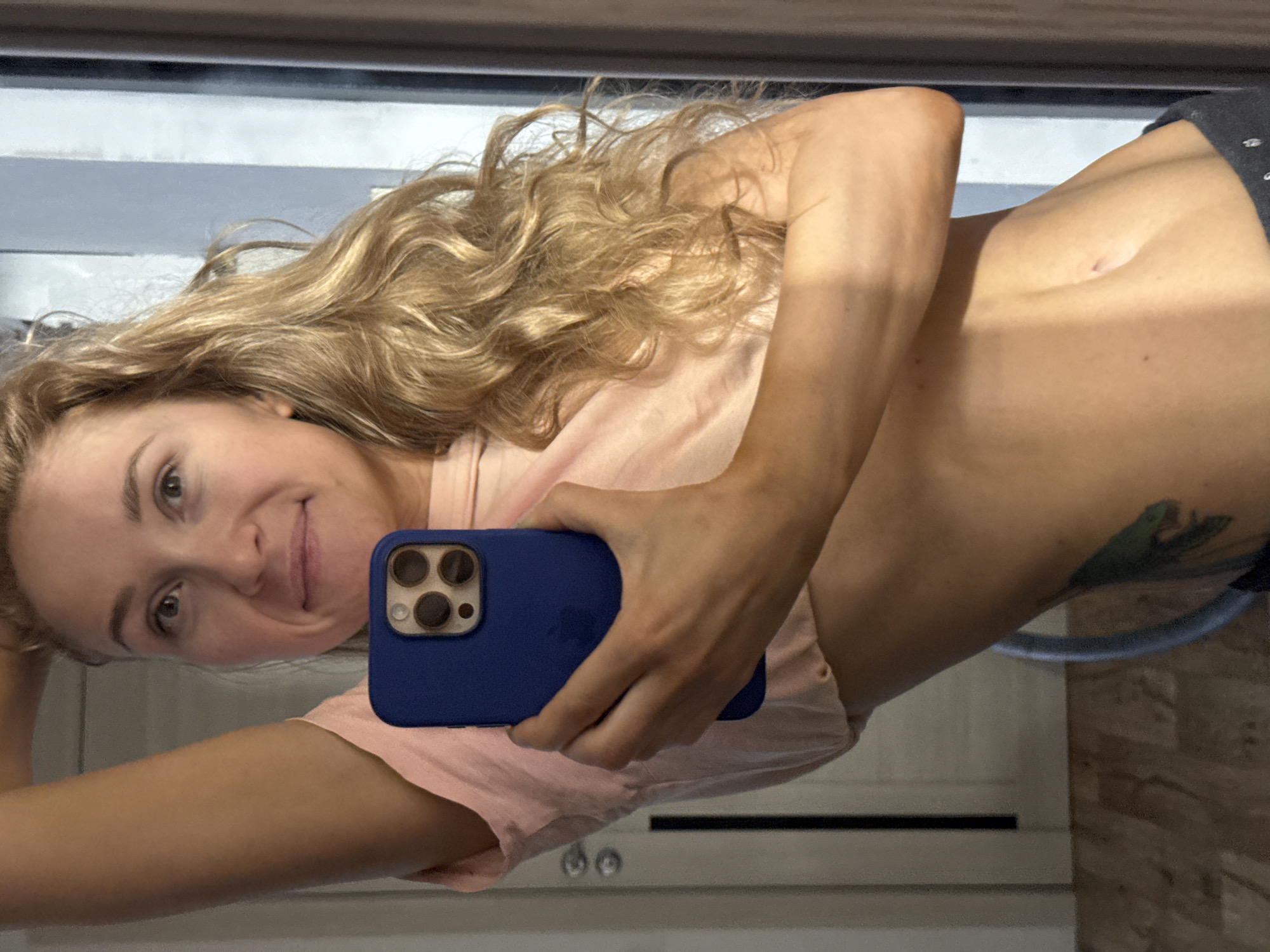 A selfie shot in a bathroom mirror shows a woman with blonde, curly hair, holding a blue-cased iPhone. She is wearing a light pink shirt, revealing her torso. A tattoo is visible on her side near her waist. The background suggests a typical bathroom setting, with wooden patterned paneling on the right-hand side of the frame and a white door in the background with silver knobs. The location of Perm, Russia is not evident within the image.