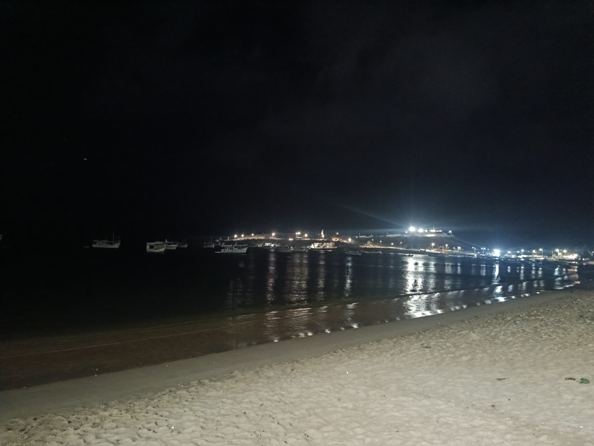 The image depicts a nighttime coastal scene in Juan Griego, Venezuela. The foreground consists of a light-colored sandy beach, showing minor textural variations and faint impressions on its surface. The beach transitions into a calm body of water, likely a bay, which occupies the midground. Several boats of varying sizes are visible on the water, appearing as dark silhouettes against the ambient light and reflections. In the distance, along the far shore, a settlement or town is illuminated by numerous bright artificial lights, presumed to be streetlights and building lights. These lights are strongly reflected on the water's surface, creating elongated streaks of illumination that extend towards the viewer. The sky above is dark, consistent with nighttime, with a single faint point of light visible in the upper left quadrant. No individuals or specific actions are discernible within the frame.