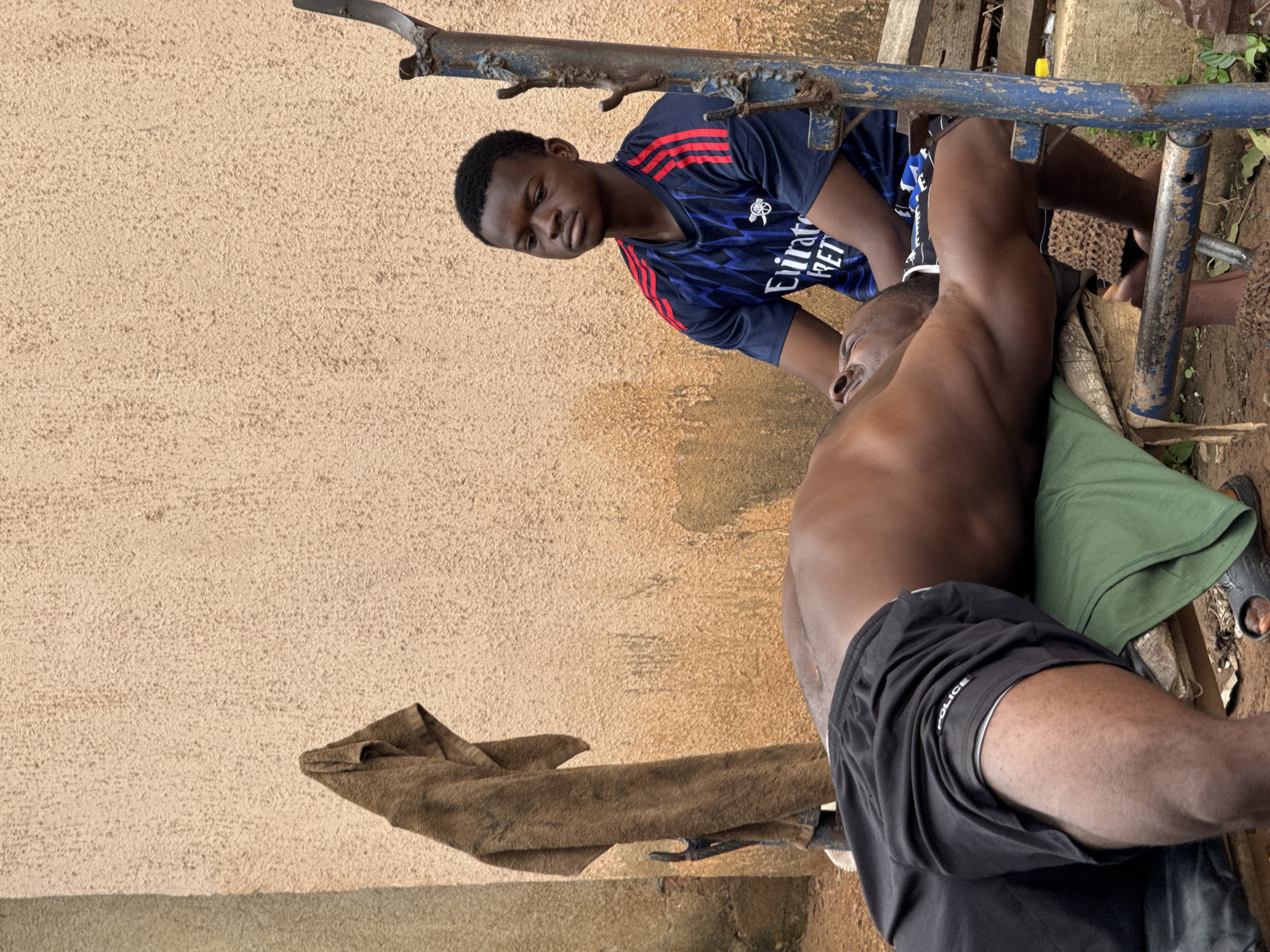 In Akwanga, Nigeria, two young men are engaged in a weightlifting activity outdoors. The man on the bench is lying on his back, lifting a weight which is not visible in the frame. The other man, seated, appears to be assisting or spotting him. The setting is rustic, with a textured, light-colored wall forming the backdrop. A brown towel hangs on a pole to the left of the frame, and a blue metal bench is utilized for the exercise. The ground is dirt and scattered with debris. The focus is on the physical exertion and the interaction between the two individuals.