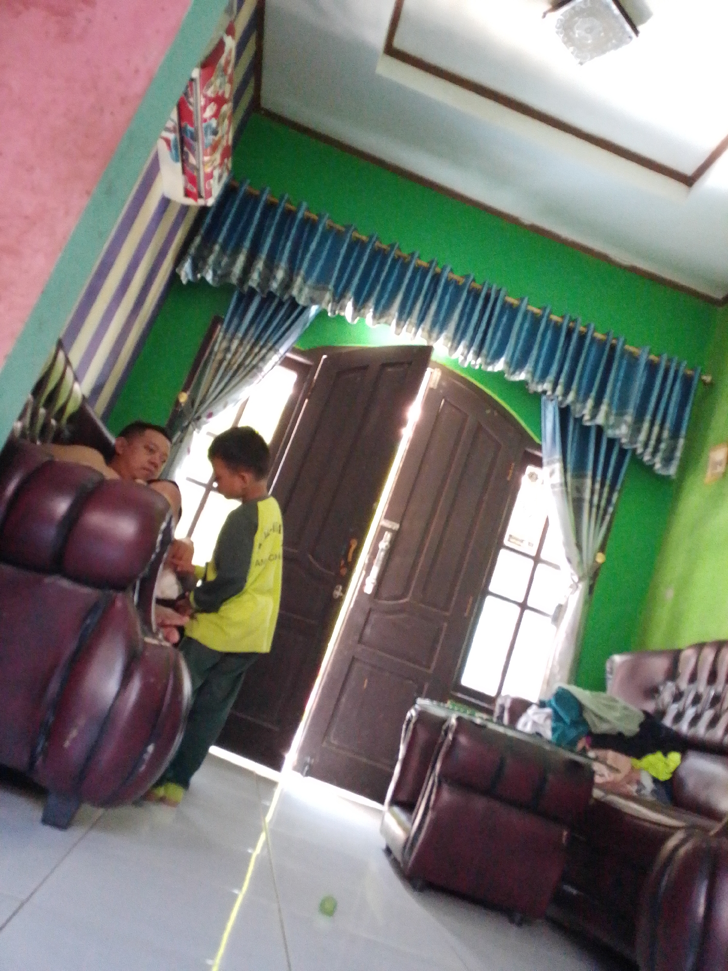 The image depicts an indoor scene, likely a living room, in a residential setting in Panyingkiran, Indonesia. The walls are painted in vivid green. A patterned curtain, in shades of blue, hangs across a window. A ceiling light fixture is visible. In the foreground, a dark red, tufted armchair is partially visible. To the left of the frame, a man, appearing to be of Asian descent, is seated. He is wearing a dark shirt and has short, dark hair. Standing to his right is a young boy, wearing a yellow long-sleeved shirt with dark sleeves and dark pants. The boy is facing the man and appears to be interacting with him, possibly reaching for something. Behind them are large double doors, one of which is slightly ajar, revealing a bright light source from outside. Further into the room, a sofa is visible, covered with various articles of clothing. The flooring appears to be tiled. The overall impression is of a casual, domestic environment.