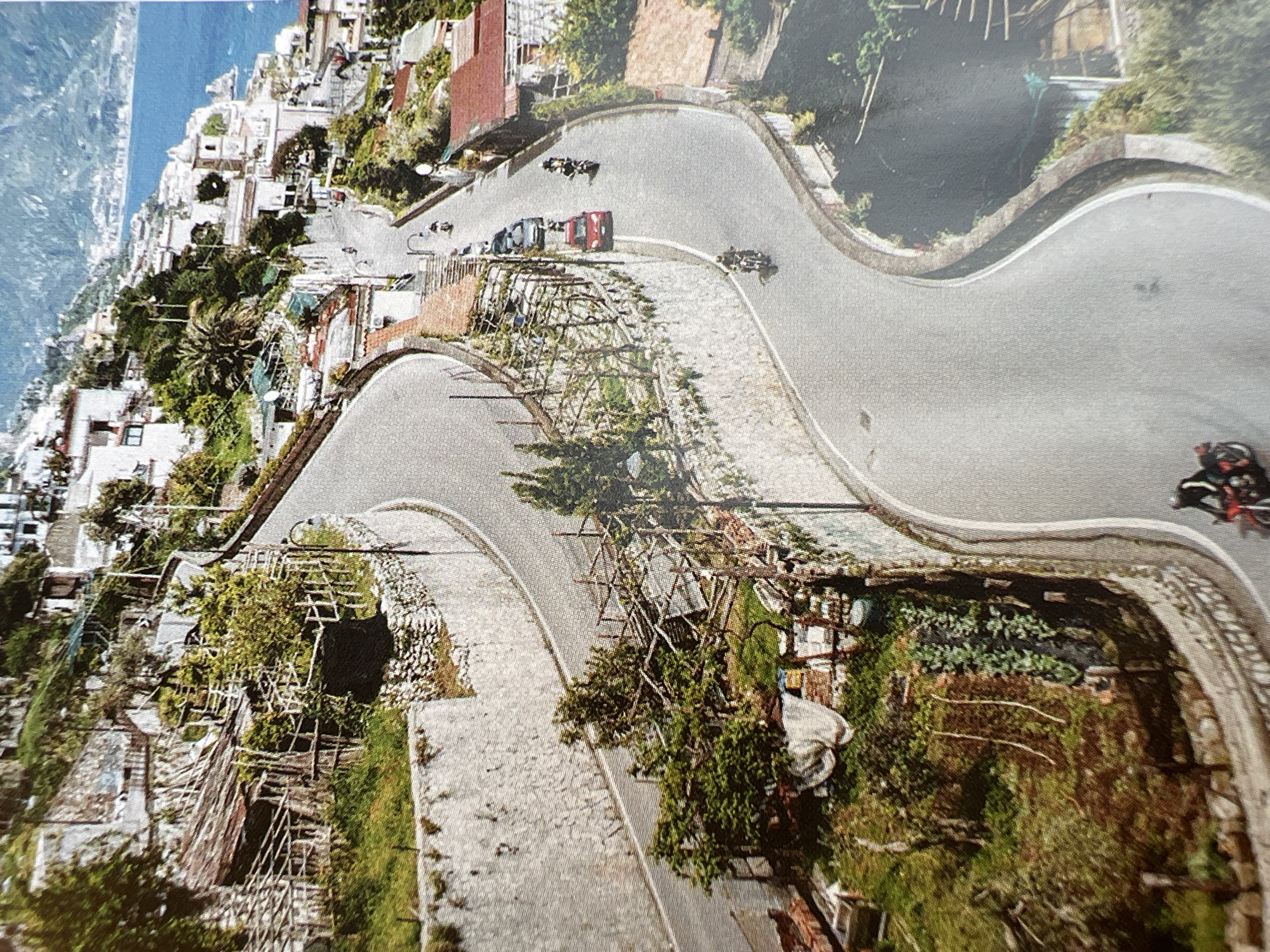 This media file depicts a scenic, winding road carved into a hillside with lush greenery and buildings clinging to the slopes. The road is paved with cobblestones in some sections and asphalt in others. Several vehicles, including cars and motorcycles, are visible on the road, suggesting a race or a tour.
In the foreground, a motorcycle with a rider is in motion. Further up the road, a red car and several other vehicles are also present. The overall scene is vibrant and bathed in natural light, suggesting it is daytime. The architecture of the buildings and the vegetation hint at a Mediterranean or similar climate. The presence of the winding road and the buildings cascading down the hillside suggest a location with dramatic topography, possibly a coastal town or a mountainous region. There is no visible text in the image.