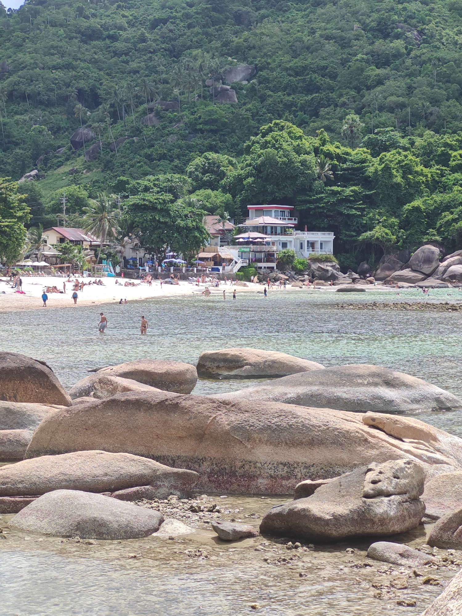 This media file captures a vibrant beach scene in Baan Chaweng Noi, Thailand. The foreground is dominated by large, rounded rocks partially submerged in clear, shallow water. The water reflects the bright daylight, creating a shimmering effect.
Beyond the rocks, a sandy beach is dotted with numerous people. Some are sunbathing on loungers, while others are walking or standing near the water's edge. A few individuals are wading in the shallow sea. The beach is lined with a collection of buildings, including what appears to be a multi-story white hotel or resort with red accents, and smaller structures with awnings, suggesting a lively tourist area. Trees, including palm trees, and lush green foliage form a dense backdrop, climbing up a steep, rocky hillside. The overall atmosphere appears relaxed and summery, indicative of a pleasant day. The time of day is likely midday, given the bright sunlight and shadows. The weather seems clear and sunny, ideal for a beach day. There is no visible text in the image.