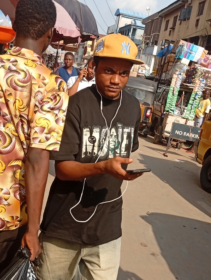 A man wearing a yellow "NY" baseball cap and a black t-shirt with an image of money, looks at the camera while holding a phone and wearing earbuds. He stands in a busy street, with pedestrians, vehicles, and makeshift stalls visible in the background. "No Parking" sign is also visible. Another man wearing a patterned shirt is partly visible from the back. The overall impression is an urban scene with the subject as the main focus.