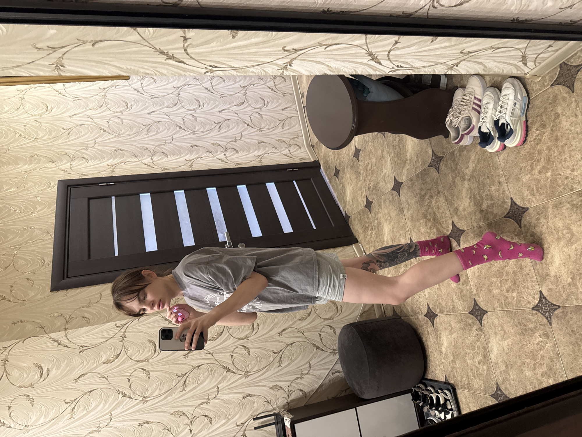 A young person is taking a selfie in a room. The individual has light skin, brown hair, and is wearing a gray t-shirt and shorts with pink socks covered in bananas. The person is holding a black smartphone and looking at the screen. In the background, there's a beige wallpaper with a floral pattern, a dark brown door with white bars, and tiled floor with a geometric pattern. There is also a grey ottoman, a shoe rack holding three pairs of athletic shoes, and a small round brown table. The scene is indoors and the image appears to be taken in a mirror. This scene is most likely from Rostov-on-Don, Russia.