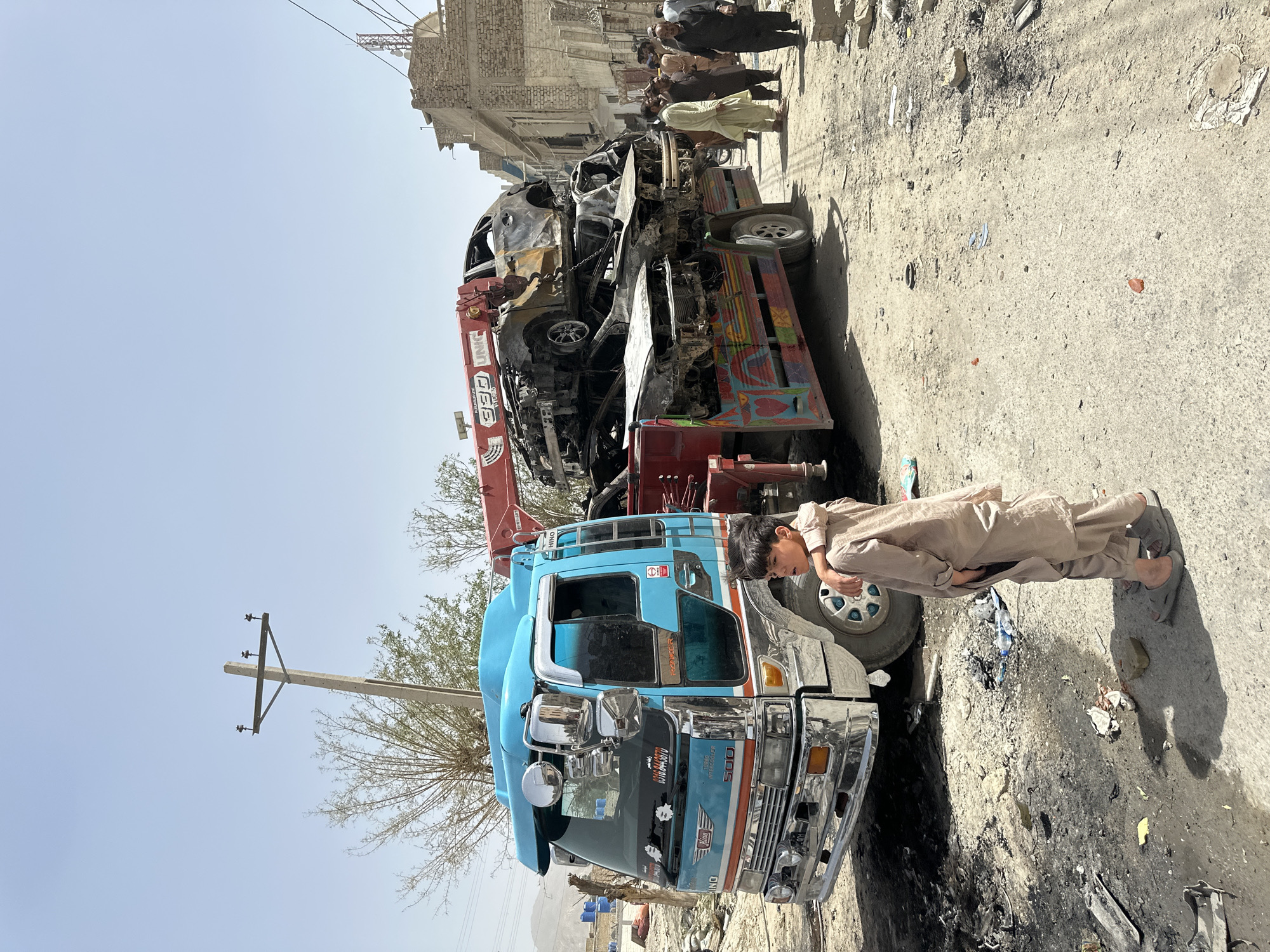 A blue Hino truck is parked on a street in Quetta City Tehsil, Pakistan. On top of the blue truck is a burnt vehicle being lifted by a red UNIC crane. A young boy in a tan outfit stands next to the truck. Several people are in the background standing in a group by a building. The street has debris scattered across it. There is a utility pole in the foreground.