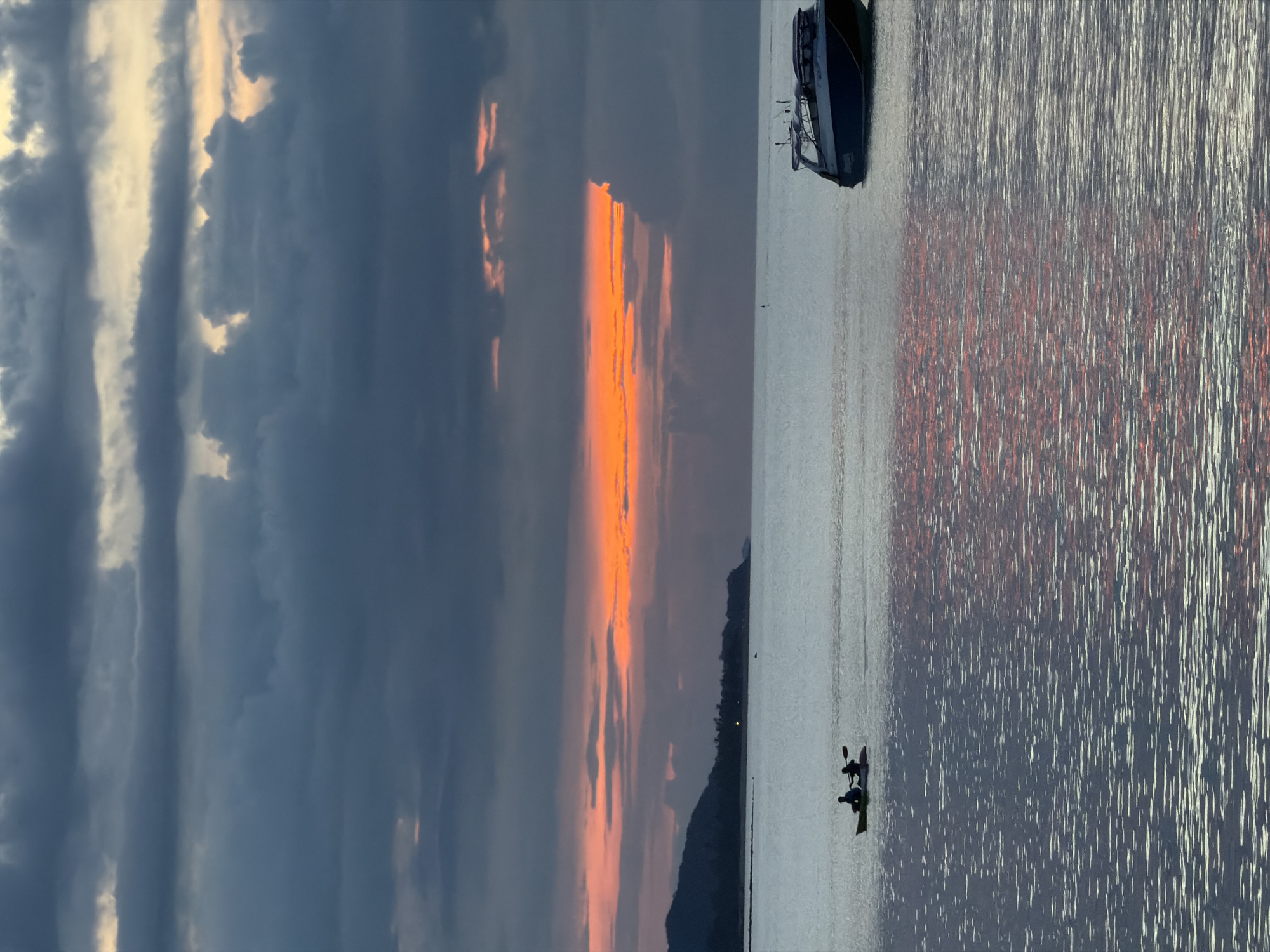 The image captures a serene seascape at dusk in Baan Bang Rak, Thailand. The sky is filled with dramatic, dark clouds, punctuated by a vibrant band of orange and yellow light near the horizon, indicating the setting sun. The sea below is calm, reflecting the colors of the sky, creating shimmering patterns.
In the middle ground, two individuals are silhouetted in a kayak, paddling across the water. To the right, a motorboat is anchored. In the far distance, a dark, mountainous silhouette of land borders the horizon, with a scattering of lights suggesting a coastal settlement. The overall atmosphere is peaceful and tranquil, with the natural beauty of the sunset dominating the scene.