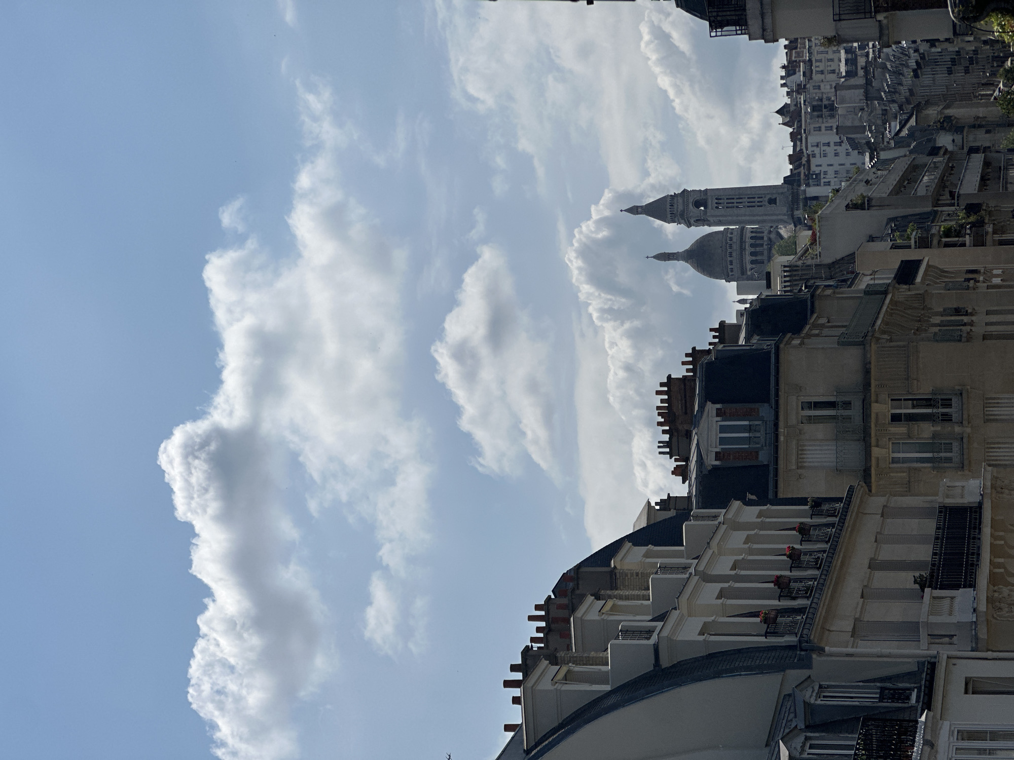 The image shows a view of Paris, France, including the Sacré-Cœur Basilica on Montmartre. The basilica's dome and tower are visible among the rooftops of the surrounding buildings. The foreground consists of Parisian architecture, with buildings featuring multiple stories, windows, and chimneys. The sky is partly cloudy with blue sky peeking through.
