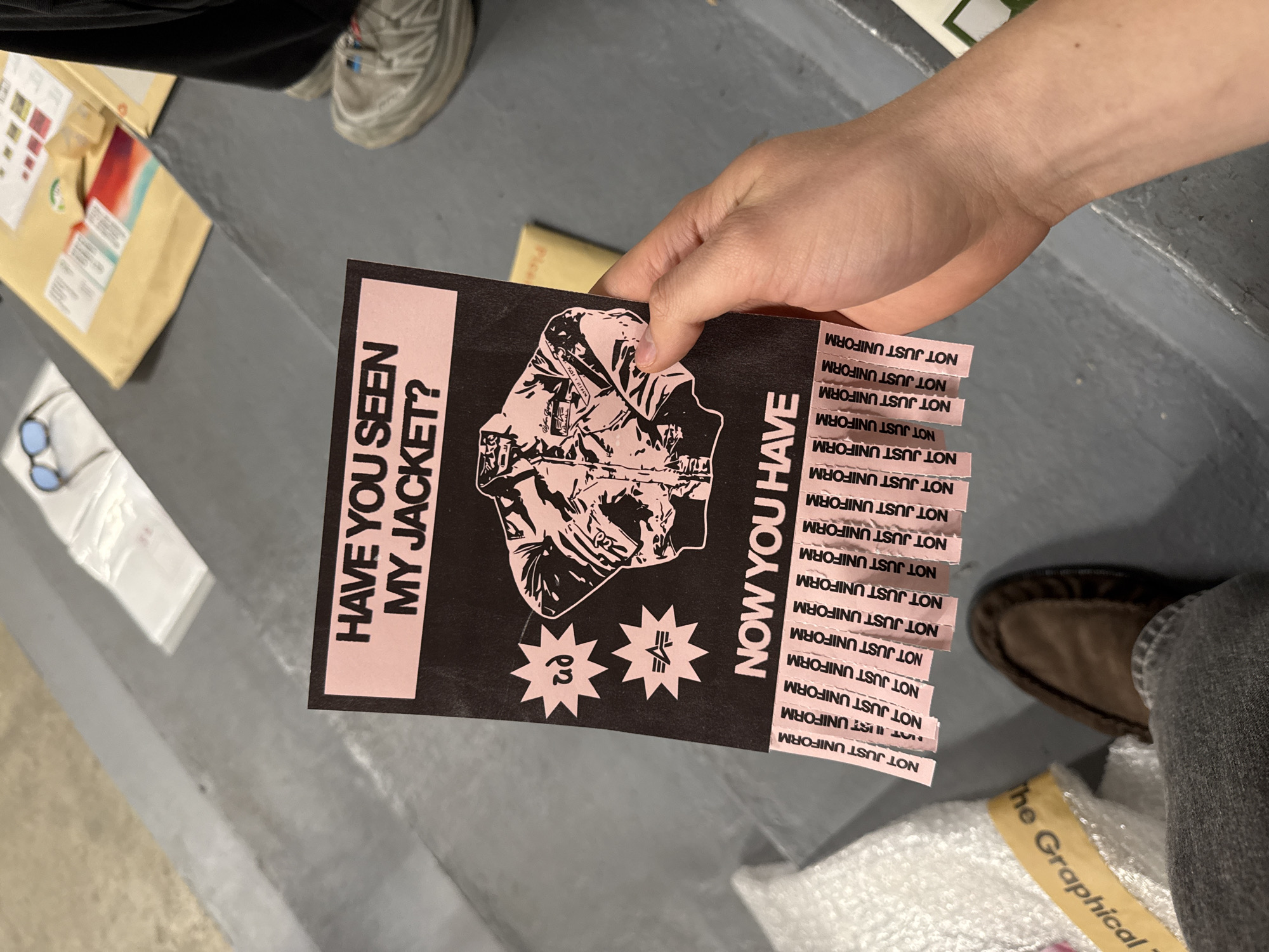 A person's hand is prominently featured, holding up a distinctive flyer designed with a black background and pink text/graphics. The flyer's left side asks, "HAVE YOU SEEN MY JACKET?", while the right side states, "NOW YOU HAVE". A detailed illustration of a jacket occupies the center. Below the jacket are two star-shaped logos; one contains the Greek letter 'ρη' and the other a stylized emblem. Along the right edge of the flyer are numerous tear-off tabs, each printed with the repeated phrase "NOT JUST UNIFORM."
The scene appears to be an indoor setting, likely a casual or creative space in London, UK. The floor is a neutral grey, possibly concrete. In the background, various items are visible: a brown cardboard parcel with labels, a pair of light blue sunglasses resting on white paper, and glimpses of two other individuals. One person is seen wearing dark pants and light-colored sneakers, while another wears grey jeans and a brown shoe. A yellow strip in the bottom right corner displays the partial text "The Graphical." The overall atmosphere is informal, suggesting the flyer is being shown or distributed.