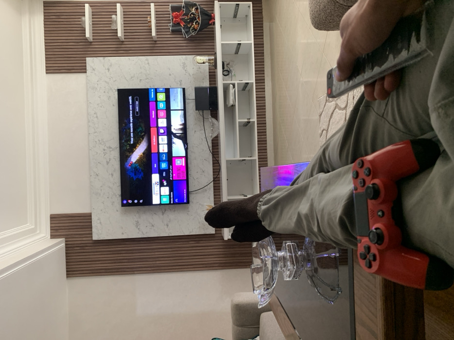 A person sits with a red PlayStation controller and a remote control in their hands, facing a TV screen displaying a user interface. Their legs are crossed, with socks on the feet resting near a glass dish on a table. The TV is mounted on a marble panel. Behind the TV, there is a bookshelf with empty shelves and decorations on the wall. The room appears to be a living space with light-colored walls and furniture.