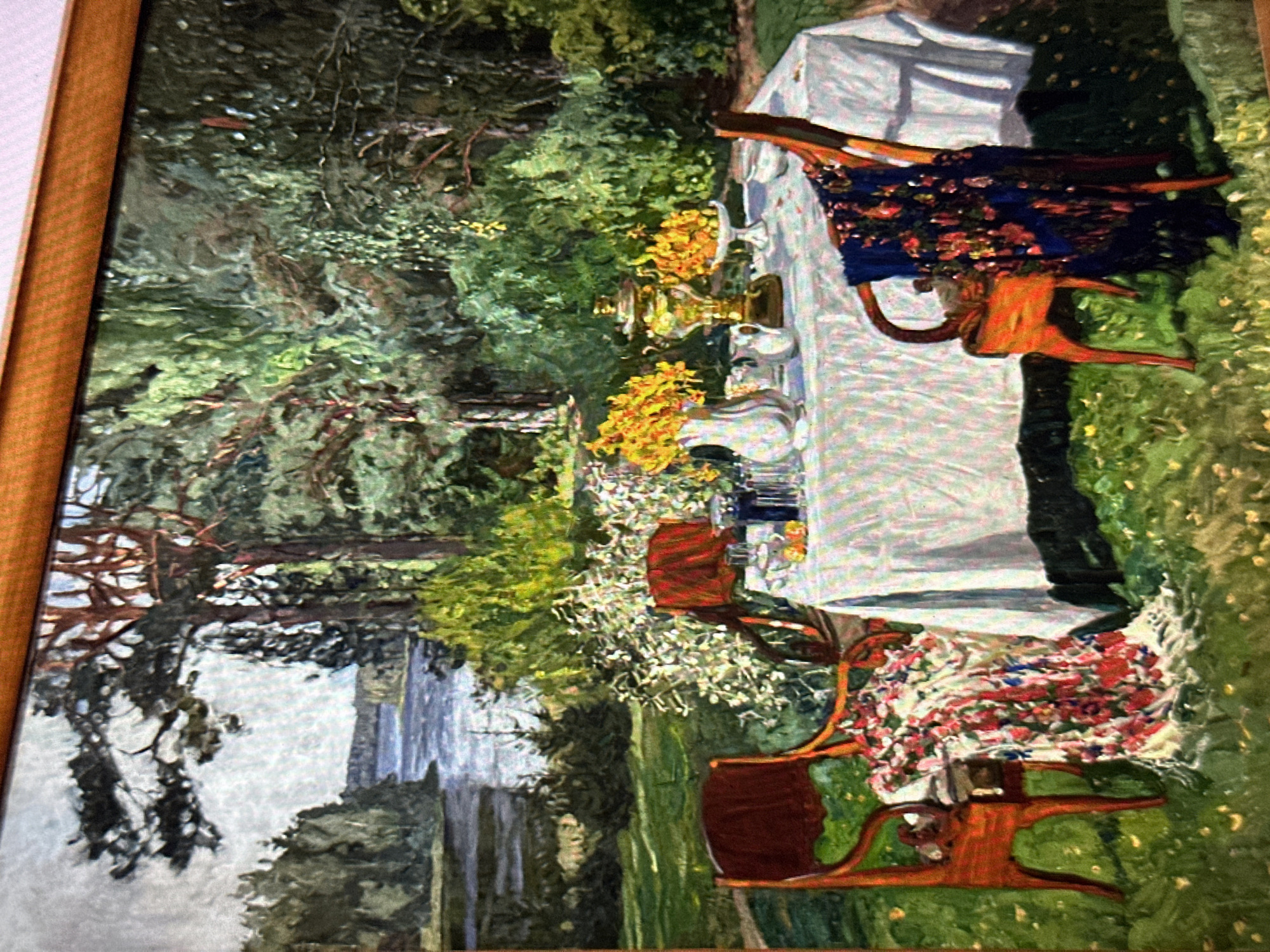 The image depicts an oil painting of a tranquil outdoor setting, likely a garden or park, near a body of water. A white tablecloth is spread over a table, laden with items for what appears to be a tea ceremony or gathering. Prominent on the table is a samovar, a traditional Russian tea urn, flanked by vases filled with yellow flowers. Several glasses and other tableware are also visible. Two wooden chairs with red upholstery are positioned at the table; one chair is draped with a colorful, floral-patterned fabric that trails onto the ground, while the other is draped with a dark blue shawl featuring a similar floral design. The background is dominated by lush greenery, with trees and bushes creating a dense forest-like environment. The presence of a lake or river in the distance suggests a natural landscape. The painting's style is Impressionistic, with visible brushstrokes contributing to a sense of light and atmosphere. This scene is not specifically located in the city of Balashikha, Russia, but rather evokes a general Russian dacha or countryside aesthetic, often associated with summer gatherings and traditional customs.
