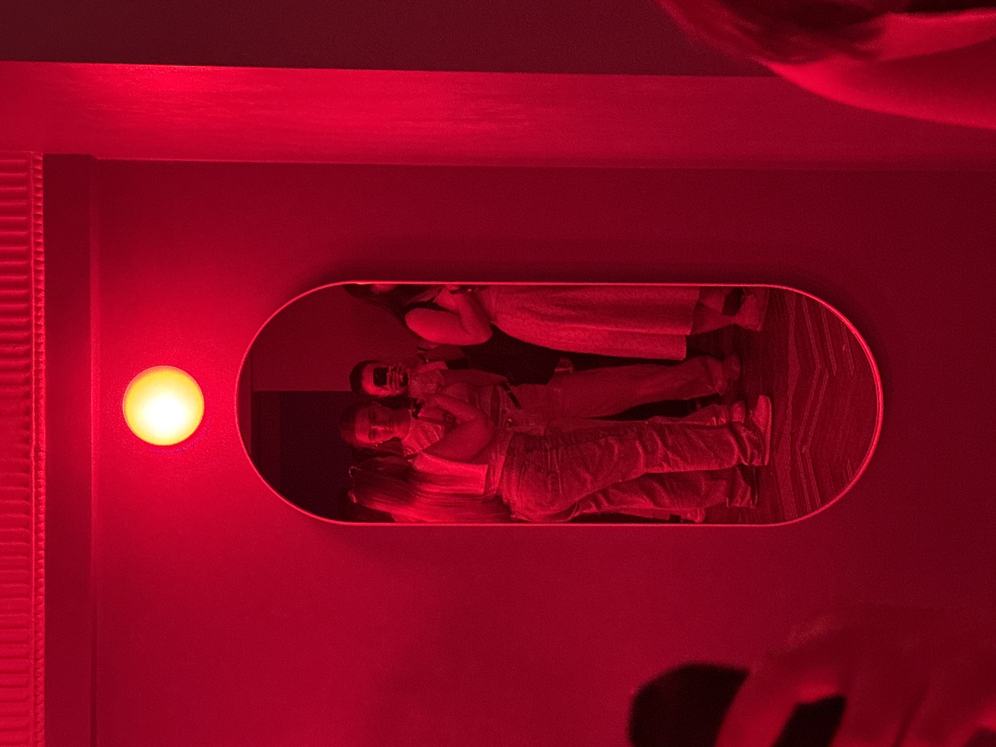 The image displays an indoor scene dominated by pervasive red lighting, emanating primarily from a single circular overhead light fixture positioned above a large, vertically oriented, oval mirror. The light fixture itself emits a bright yellow-orange glow, illuminating the surrounding area in a deep red hue.
The mirror, centrally located on a plain red wall, reflects several individuals. On the left side of the reflection, a person with long hair is seen from the back, wearing a light-colored crop top and light-wash jeans, facing another individual. This second person, positioned to the right of the first within the mirror, is holding a black smartphone up to their face, seemingly taking a photograph or video. Portions of their upper body and face are visible. The lower extremities of at least one other person are visible at the bottom of the mirror's reflection, wearing light-colored shoes.
On the right side of the mirror's reflection, another individual is partially visible from the side/back, wearing a light-colored, patterned dress that extends below the knee. Their dark hair is visible. Further to the right in the reflection, an arm and torso suggest the presence of another person. The reflected floor appears dark with subtle patterns.
The immediate environment outside the mirror includes a red wall. Above the mirror, decorative molding or a cornice runs along the ceiling line, also appearing red. To the right of the mirror, a red-lit architectural feature, possibly a column or wall corner, is visible. A blurry, dark shape in the bottom left foreground of the original image, outside the mirror's frame, indicates an object or part of another person close to the camera. The scene lacks specific visual cues to confirm the geographical location as Укромновское сельское поселение, Ukraine.