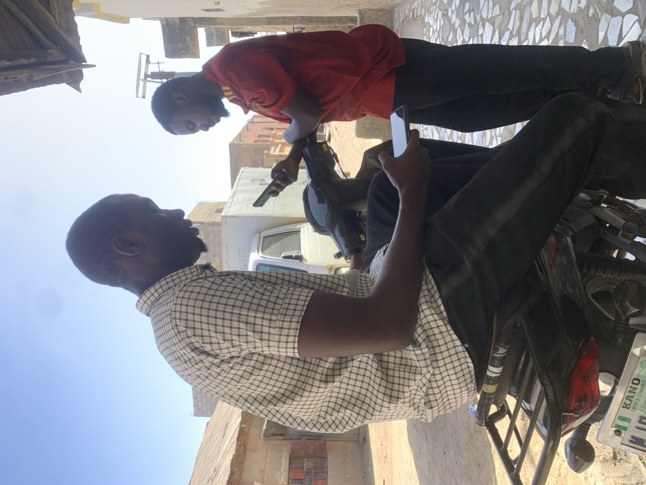 Two adult males are depicted outdoors in Gwagwarwa, Nigeria, under clear daylight. The male on the left, wearing a white and black checkered short-sleeve shirt and dark pants, is seated on a dark motorcycle. He holds a white smartphone horizontally in his left hand, looking downwards at its screen. The motorcycle has a black seat, a rear rack, and a partially visible Nigerian license plate from "KANO CENTRE OF COMMERCE." The male on the right, wearing a red short-sleeve shirt and dark pants, stands alongside the motorcycle. He holds a dark smartphone vertically with both hands, focusing on its screen. In the background, a white boxy vehicle is parked near several buildings, including one constructed from concrete blocks and another with a corrugated metal roof. The ground surface in the immediate foreground right is composed of irregular light-colored paving.