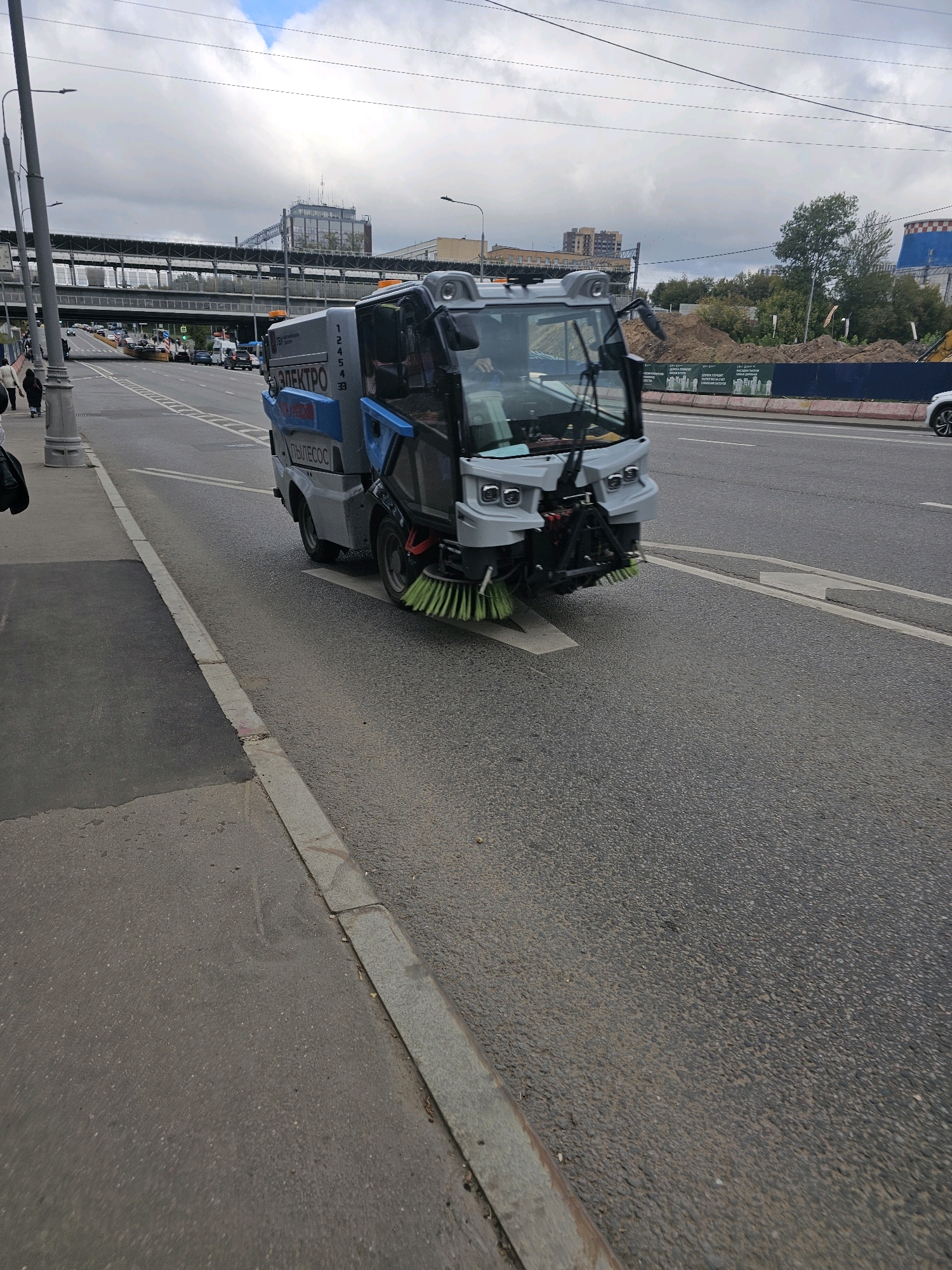 A street sweeper vehicle is shown on a paved road in Moscow, Russia. The street sweeper, which is predominantly grey and blue, has its brushes extended and appears to be in operation. The vehicle has the Cyrillic text "ЭЛЕКТРО" and "ПЫЛЕСОС" visible on its side. The road is asphalt, with a concrete curb and sidewalk on the left. In the background, there is a multi-level overpass, several buildings, trees, and utility poles. The sky is overcast with clouds. Several cars are visible on the road in the distance, and a few pedestrians are on the sidewalk. The overall environment suggests an urban setting.