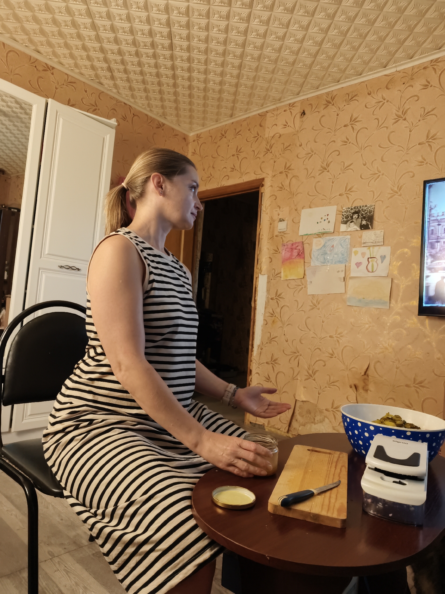A young woman with blonde hair pulled back in a ponytail, wearing a black and white striped sleeveless dress, sits at a round wooden table in what appears to be a dimly lit room. She is facing to her right, looking towards a television screen visible in the upper right corner of the frame. Her left hand is resting on the table, near a small jar with a yellow lid and a wooden cutting board with a knife. Her right hand is extended outwards, palms up, in a gesturing motion.

On the table, in addition to the jar, cutting board, and knife, there is a white electric appliance with a black top and a blue and white polka dot bowl filled with chopped pickles. The walls of the room are covered with textured, peach-colored wallpaper featuring a repeating floral pattern. A doorway is visible behind the woman, leading into another room with a darker interior. The ceiling is covered with beige, textured tiles. Several children's drawings and photographs are affixed to the wallpaper on the wall to the right of the doorway. The room appears to be in a residential setting. The image does not contain specific visual cues to confirm the location within the city of Star, Russia.