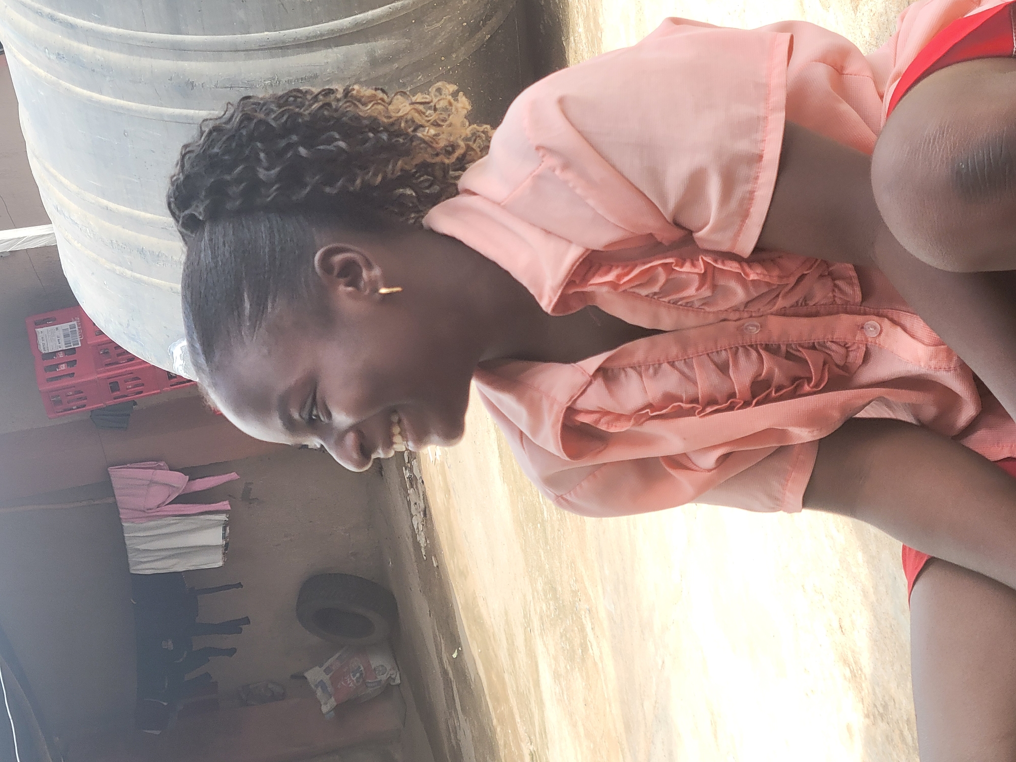 A young woman is outside, likely in a residential area of Madalla, Nigeria, based on the presence of a water storage tank, discarded tire, and laundry line. The woman, possibly a teenager, has a shaved head with curly hair extensions. She's wearing an peach colored frilly button up shirt with red shorts. She is crouching. The environment includes a water storage tank, laundry hanging on a clothesline, a tire, and general debris. The foreground shows sunlit pavement or ground.