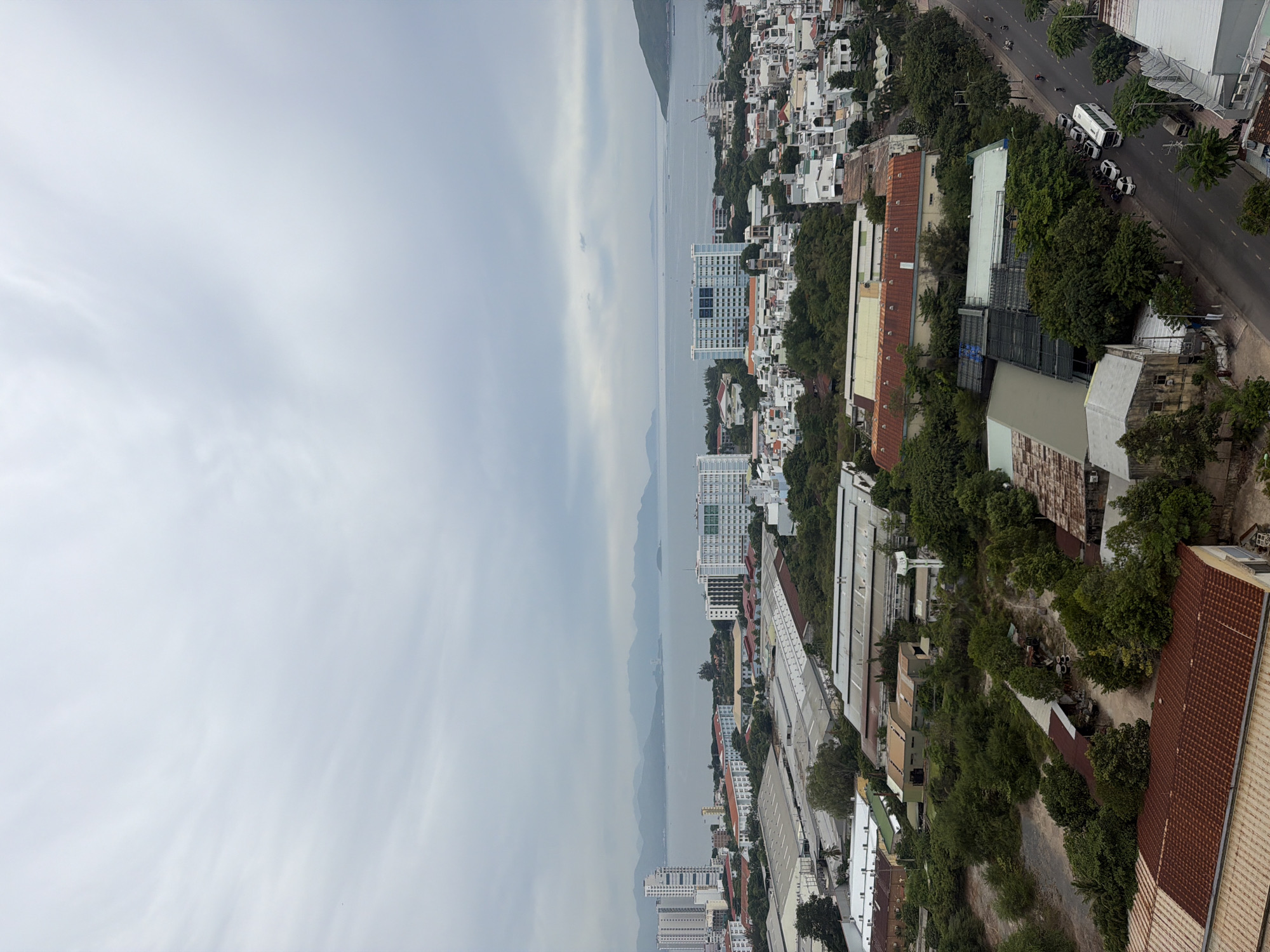 The image depicts a panoramic view of the city of Nha Trang, Vietnam, with a body of water and mountainous terrain in the background. The scene is characterized by a mixture of residential buildings, industrial structures, and lush greenery, all under a cloudy sky. In the foreground, a street is visible with several vehicles, including cars and a bus, suggesting ongoing urban activity. The buildings in the city range from modern high-rises to more traditional, lower-rise structures, interspersed with trees and vegetation, creating a sense of a densely populated yet green urban environment. The water in the background appears to be a bay or ocean, with faint outlines of islands or peninsulas on the horizon. The overall impression is that of a coastal city on an overcast day.
