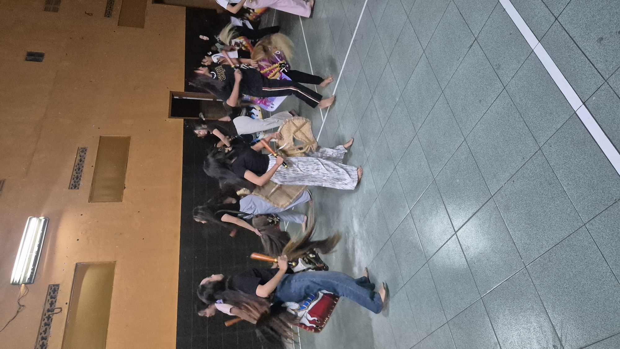 In a brightly lit indoor space with a speckled grey tiled floor and an orange-painted wall, several young women are engaged in a performance. The women, all appearing to be teenagers or young adults, are dressed casually. Some wear black t-shirts and jeans or patterned trousers, while others are in lighter colored attire. They are holding and interacting with traditional Indonesian props that resemble horse-like figures, possibly for a Jaranan performance. These props are made of wood and decorated with colorful fabrics and horsehair. The women are in motion, suggesting they are dancing or parading. The room appears to be a gymnasium or multipurpose hall, with fluorescent lighting fixtures visible on the ceiling. The overall atmosphere suggests a cultural event or practice session taking place in Sumowono, Indonesia.