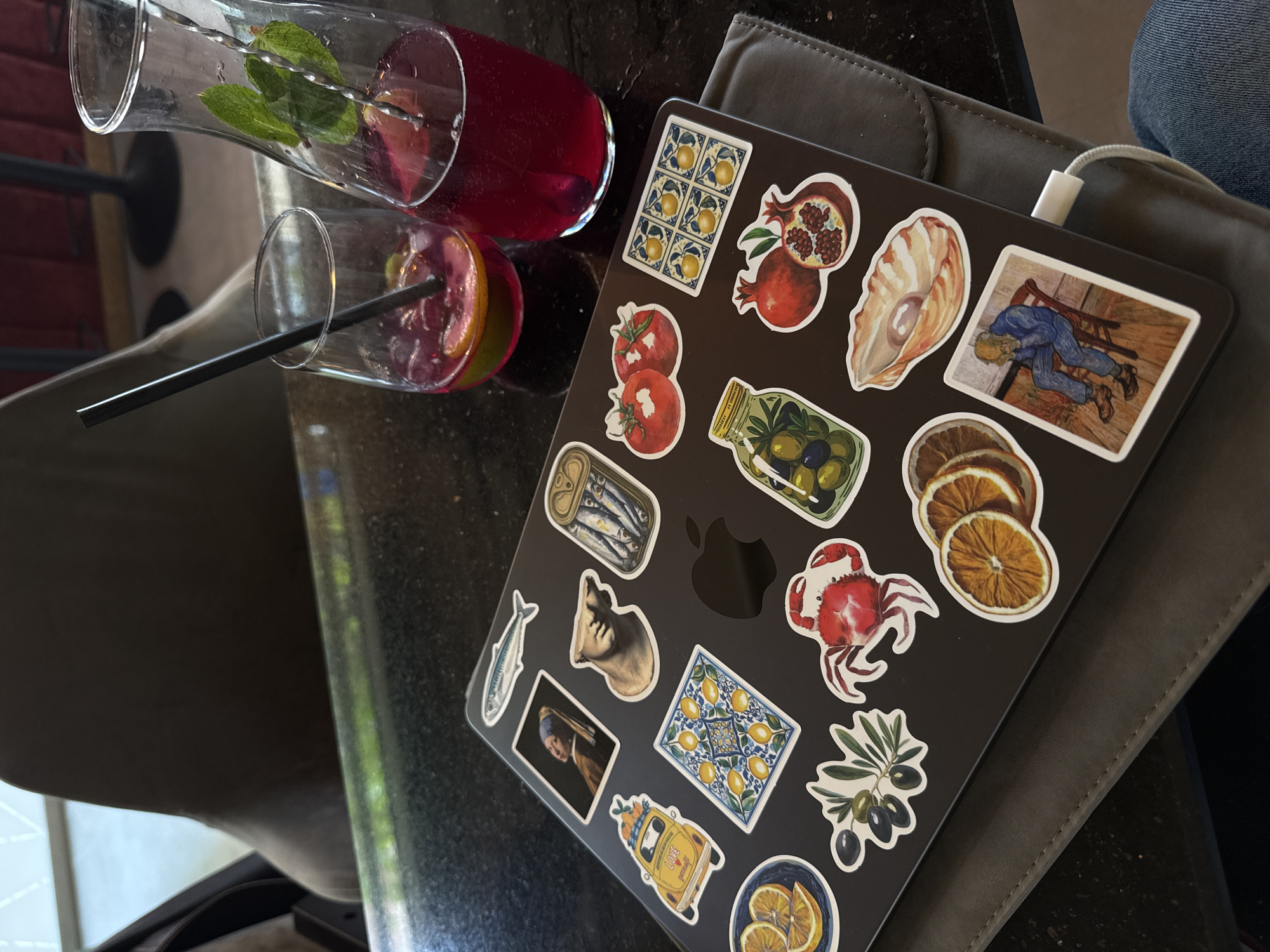 The image captures a close-up, overhead view of a black laptop adorned with numerous art and food-themed stickers, placed on a dark table surface. Beside the laptop are two glasses and a pitcher containing a vibrant red or pink beverage, garnished with fresh mint leaves and citrus slices. One glass has a black straw.
The laptop's lid is covered with a diverse collection of stickers, including: two tiled patterns featuring yellow lemons, two pomegranates (one cut open), two red tomatoes, a jar of green and black olives, a seashell, a can of sardines, a silver fish, a classical bust, a sprig of olives, a red crab, and dried orange slices. Also prominent are stickers depicting famous artworks: "Girl with a Pearl Earring" and a scene resembling Vincent van Gogh's "The Prisoner's Round," focusing on a figure hunched over a chair. A yellow vintage car sticker, with "LOVE" written on its side, is also visible. The Apple logo of the laptop is visible beneath some stickers.
The setting appears to be an indoor or shaded outdoor area, with a dark, possibly granite or marble, table. A grey laptop sleeve or case is partly visible to the right of the laptop, along with a portion of blue jeans, suggesting a person is seated nearby. A white charging cable is plugged into the laptop. The drinks are likely refreshing, given the mint and fruit garnishes.