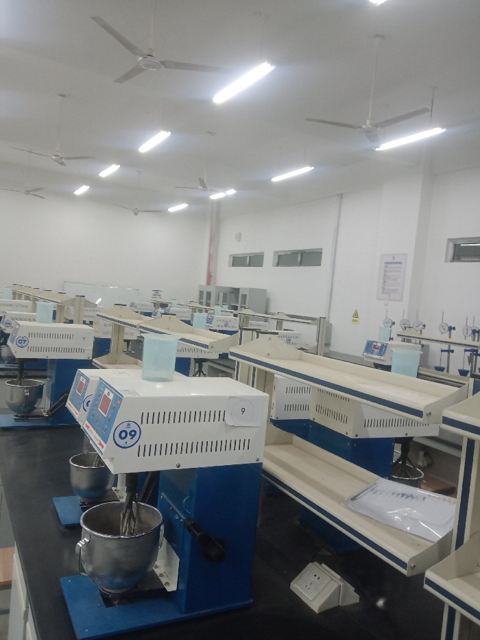 An interior view shows a well-lit, sterile laboratory setting. Rows of blue and white testing equipment with stainless steel mixing bowls dominate the foreground, marked with numbers '07' and '09'. Behind them, a long countertop features additional testing stations. Fluorescent lights and ceiling fans illuminate the space, with windows along the back wall. A red pipe runs vertically near the corner. Overall, it is a functional, orderly, and professional workspace.