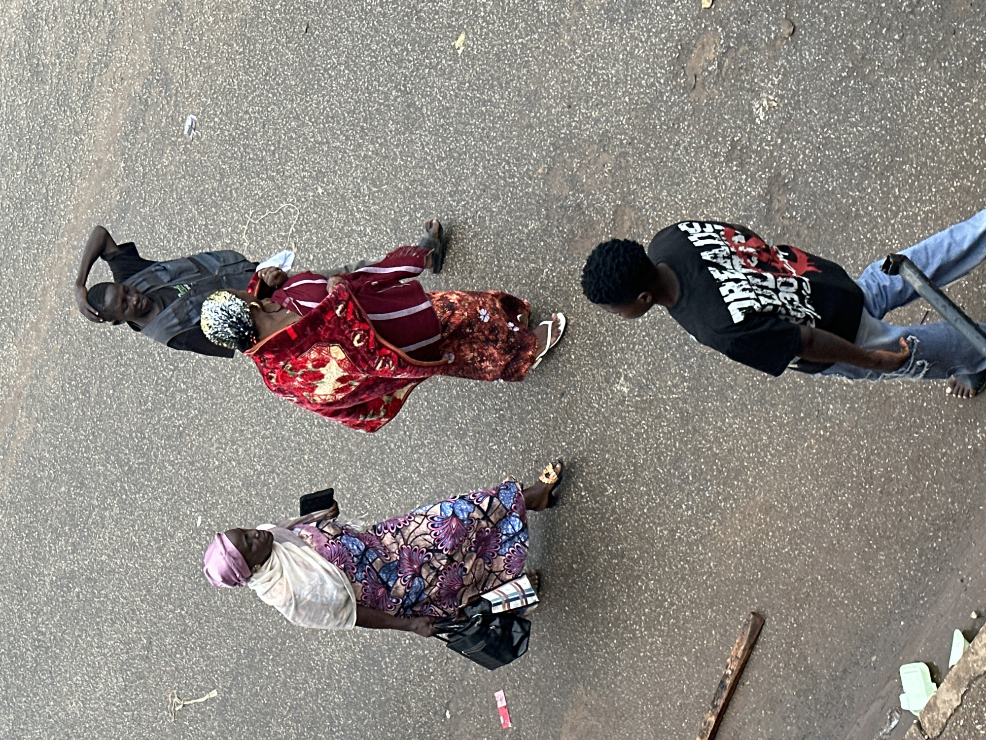 In Bukuru, Nigeria, four people are visible on a textured, asphalt surface. In the foreground, a young male with dark, curly hair wears a black t-shirt with red and white graphic text and light blue jeans. He is walking away from the camera, holding a dark metal pole. Behind him and slightly to the left, a woman with a pink headscarf and a floral patterned dress, carrying two bags, walks to the left. To her right, a woman in a red patterned garment is walking forward, and a man in a dark vest over a t-shirt and dark pants is standing with his right hand on his head. The environment is a street with scattered debris including a piece of wood and some plastic items.
