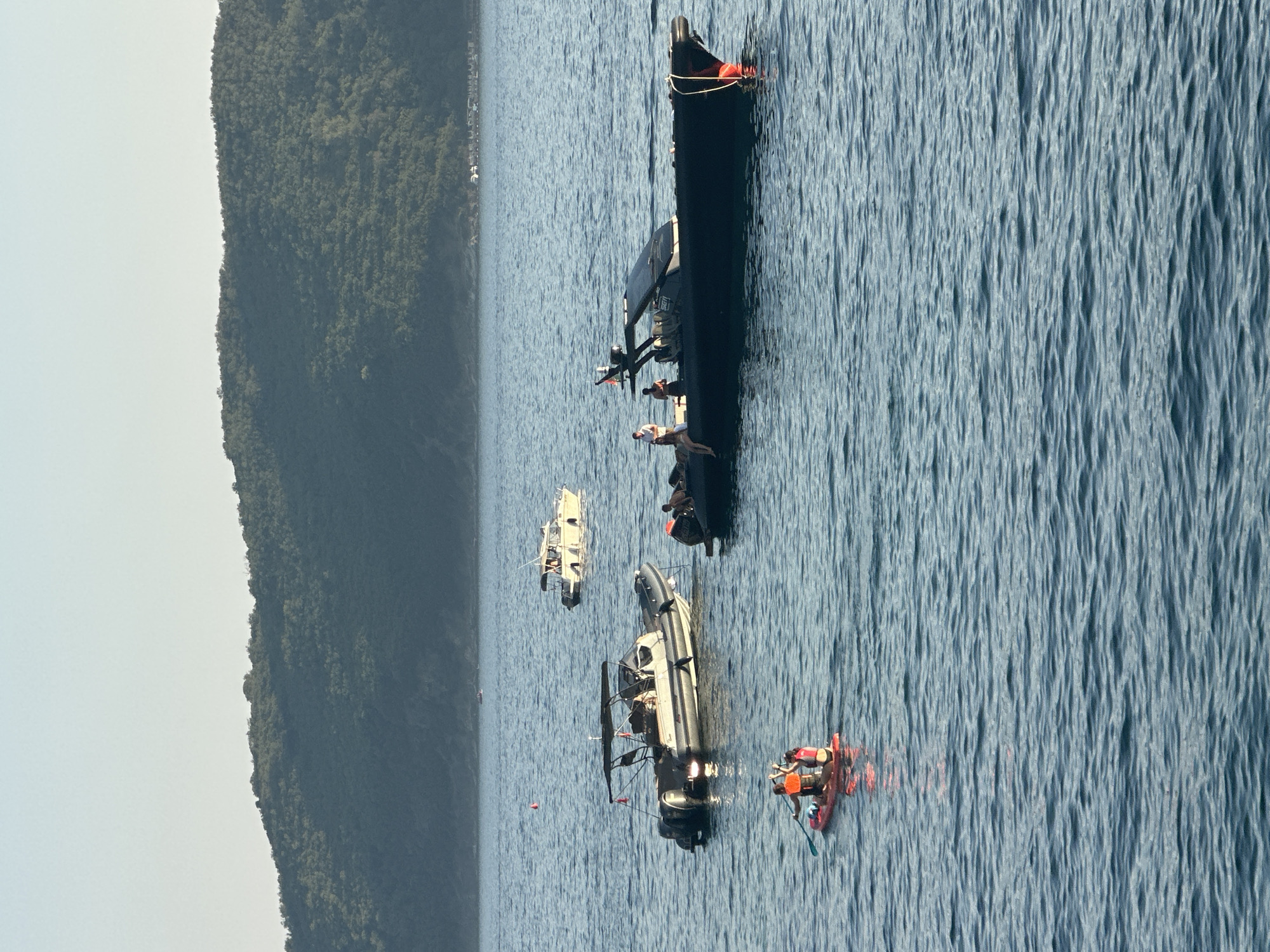 The image shows a body of water with several boats and two people on a stand-up paddleboard. In the foreground, two people are on a red stand-up paddleboard, one is paddling and the other is sitting. Behind them, a gray inflatable boat with a canopy is anchored. To the right of the inflatable boat is a black, sleek motorboat with several people standing and sitting on it. In the background, a white boat is moving away from the viewer. Further in the distance, a forested hill slopes down to the water. The water is blue and rippled, and the sky is a clear, pale blue. This scene is characteristic of the coastline near Budva, Montenegro, likely during daylight hours.