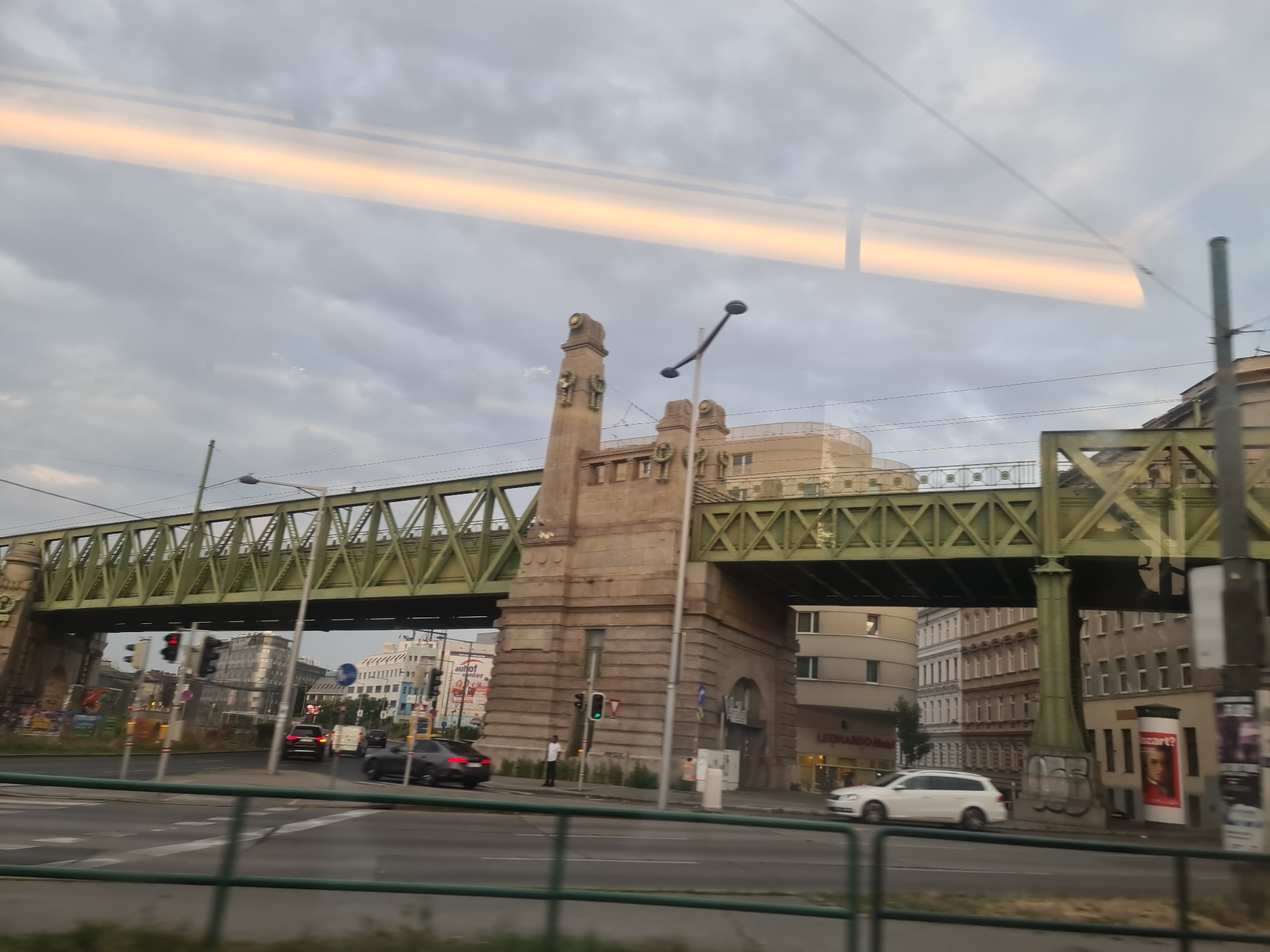 This is a photograph of the Gürtel in Vienna, Austria, taken from a moving vehicle. The image is dominated by a large, green, steel railway bridge that spans across a street. The bridge is supported by a substantial stone structure on the left side of the frame. Several buildings are visible behind and under the bridge, some appearing to be apartment buildings or offices. A white station wagon is parked on the right side of the street. Traffic lights are visible on the street, indicating ongoing traffic flow. The sky is overcast with a band of warm light, possibly from a sunset or sunrise, reflecting on the surface of what appears to be a window, creating horizontal bands of light across the upper portion of the image. The overall impression is of an urban environment with elevated infrastructure.