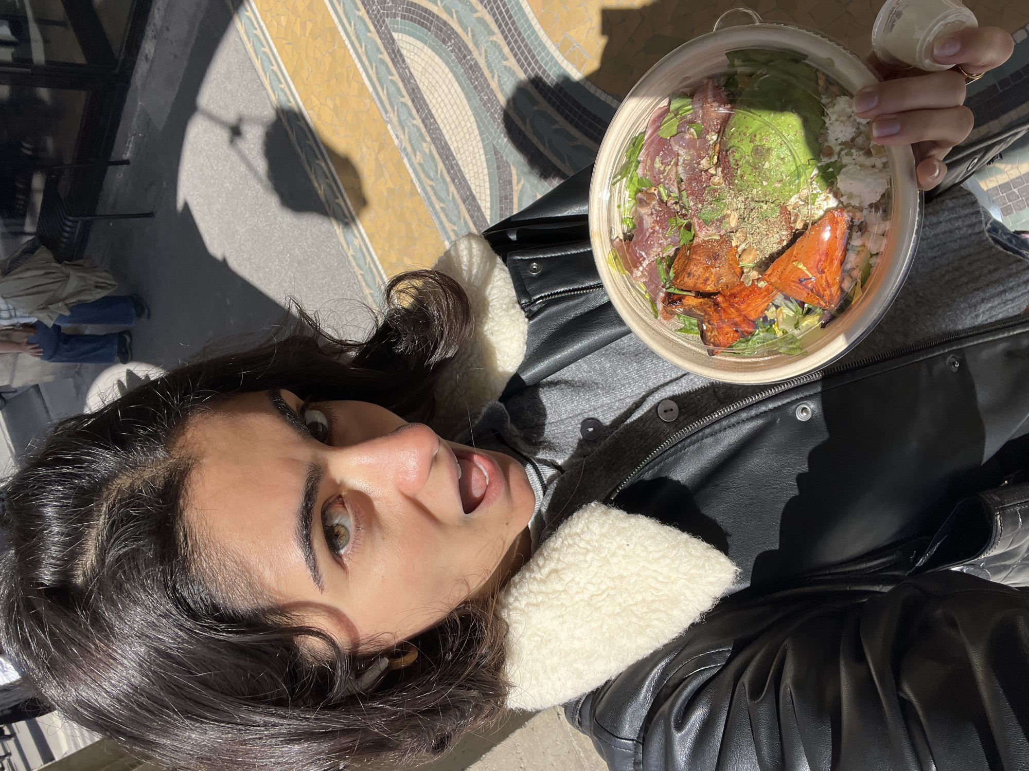 A woman takes a selfie while holding a bowl of food, possibly salad, in the city of Paris, France. The woman is wearing a grey shirt and a black leather jacket with a white wool lining. She has dark curly hair, fair skin, and wide eyes. Her mouth is open in a surprised expression. The bowl of food appears to contain lettuce, avocado, some red meat or vegetables, and cheese. The background shows a mosaic floor, with shadows falling across it. There is also a wrought iron railing visible in the background.