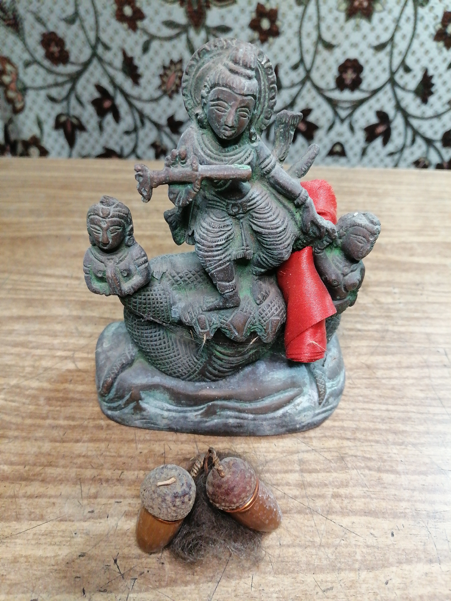 A close-up, eye-level shot features a small, weathered bronze statue of a deity playing a flute, seated on a coiled serpent. Two smaller figures are positioned on either side of the serpent. A piece of red fabric is draped over the deity's right shoulder. The statue rests on a flat, wooden surface with visible grain and scratches. In the foreground, two acorns, one with a visible stem and surrounded by dark, fibrous material, lie on the same wooden surface. The background consists of a patterned textile with a floral design in muted colors. The image is not identifiable as being in Rostov-on-Don, Russia, or any specific city.