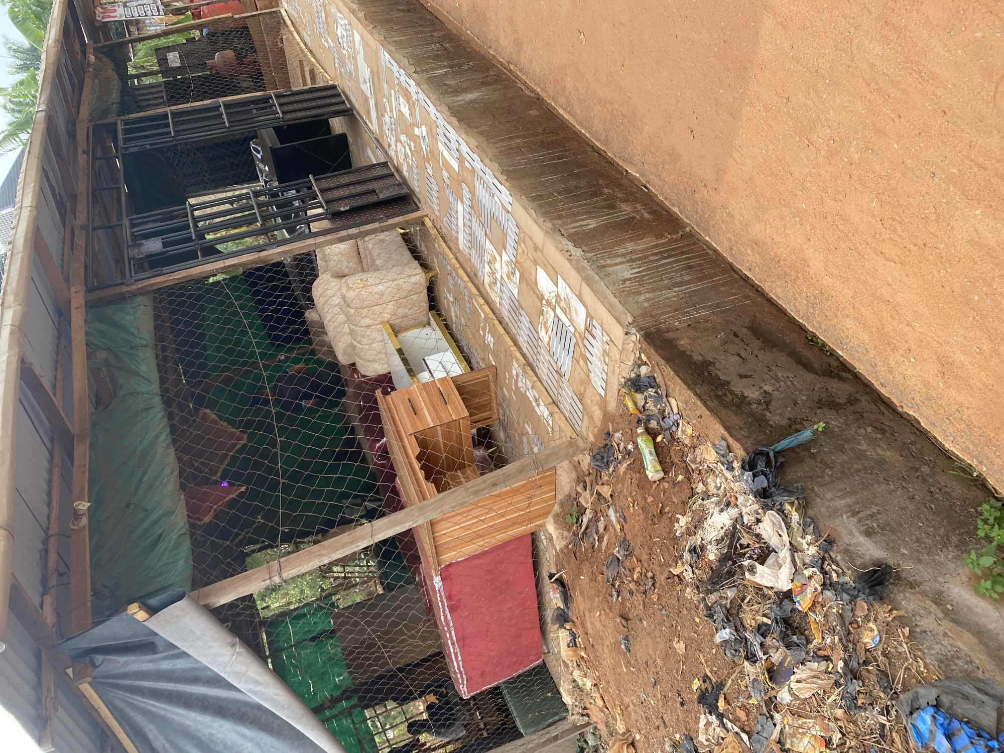 The image depicts an outdoor commercial setting in Nsukka, Nigeria. A makeshift shop structure occupies the midground, fronted by a heavily littered dirt area and a concrete drainage ditch, with a reddish-brown dirt road beyond.
The shop features a roof constructed from corrugated metal sheets supported by wooden beams, with sections of dark and green tarpaulin visible beneath. Its enclosure is formed by chain-link fencing, partially covered by green tarpaulin, and a dark grey tarpaulin is rolled up on the far left. An open black metal gate serves as the entrance.
Inside the shop, a selection of furniture is visible: several light-colored, upholstered sofas with patterned fabric and gold-colored accents, a red upholstered furniture piece, a light-colored wooden desk with drawers, and a white and wood console table. A red gas cylinder stands near the right side of the entrance, and a white sign displaying "POS" is attached nearby. A light fixture hangs from the roof structure. Two individuals are partially visible in the far left background, obscured by the fencing and merchandise.
The immediate foreground on the left consists of reddish-brown earth heavily covered with refuse, including numerous black and white plastic bags, wrappers, and debris. This area abuts a concrete drainage channel with stained walls, containing some small green plant growth at its base. Adjacent to the channel is a raised concrete walkway with a light-colored, patterned tiled surface that exhibits wear and damage. To the right of the ditch and walkway is a reddish-brown dirt road. Green foliage and trees are visible in the background behind the shop structure.