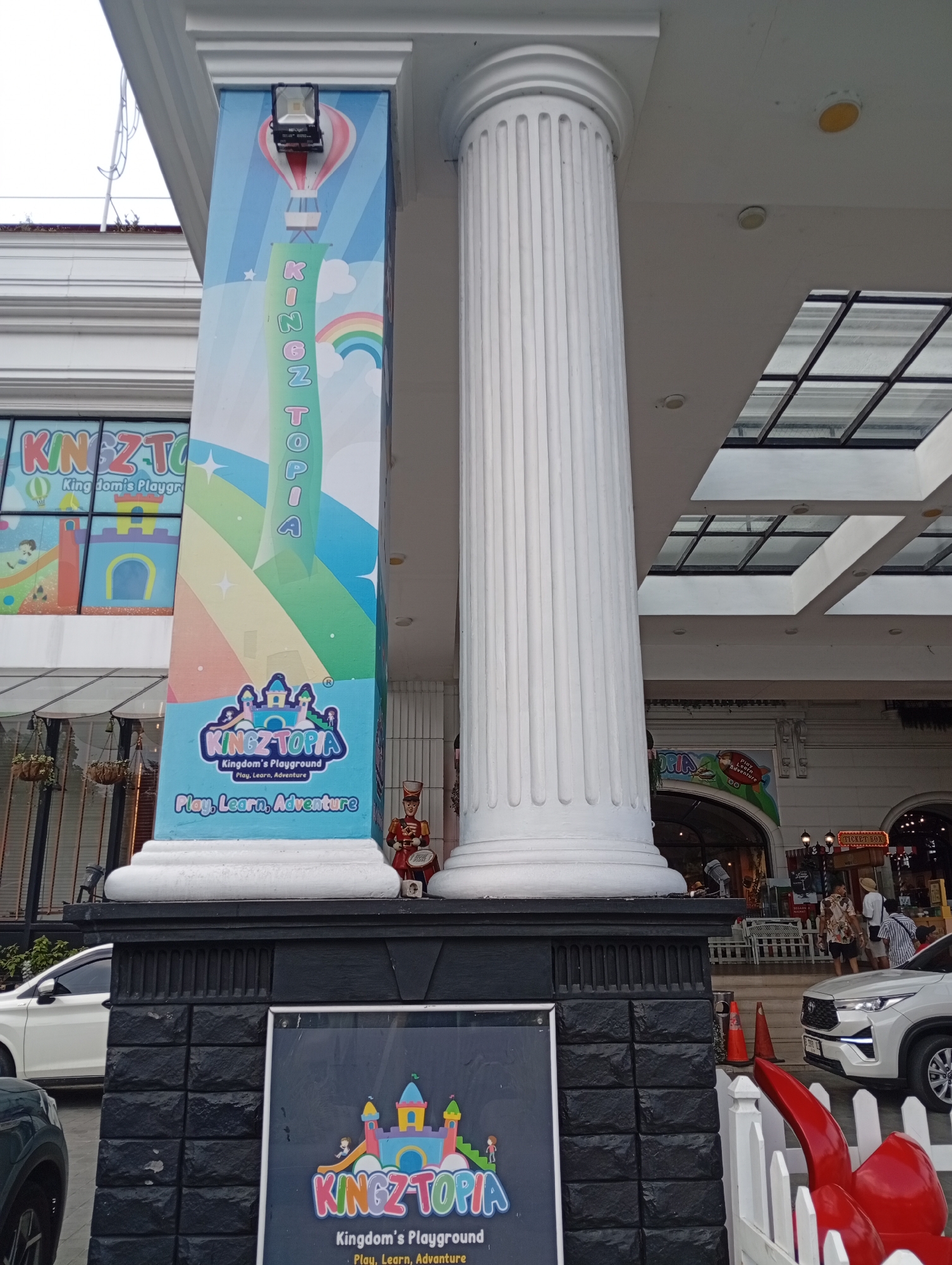The image shows the entrance to "Kingztopia," a children's playground located in Cimacan, Indonesia. A large, fluted white column stands prominently in the foreground, partially obscuring a brightly colored pillar adorned with the Kingztopia logo, a hot air balloon, and a rainbow. The pillar also features the words "Kingztopia" vertically. Below the pillar, a dark, textured base supports a sign with the Kingztopia logo and slogan: "Kingdom's Playground - Play, Learn, Adventure." A small, costumed figure, possibly a toy soldier, stands near the base of the pillar.
In the background, part of the building's facade is visible, including a glass-paneled roof structure. To the left, a white car is parked. To the right, people are seen entering what appears to be a mall or entertainment complex. The overall scene suggests a bright, family-friendly atmosphere associated with the playground.